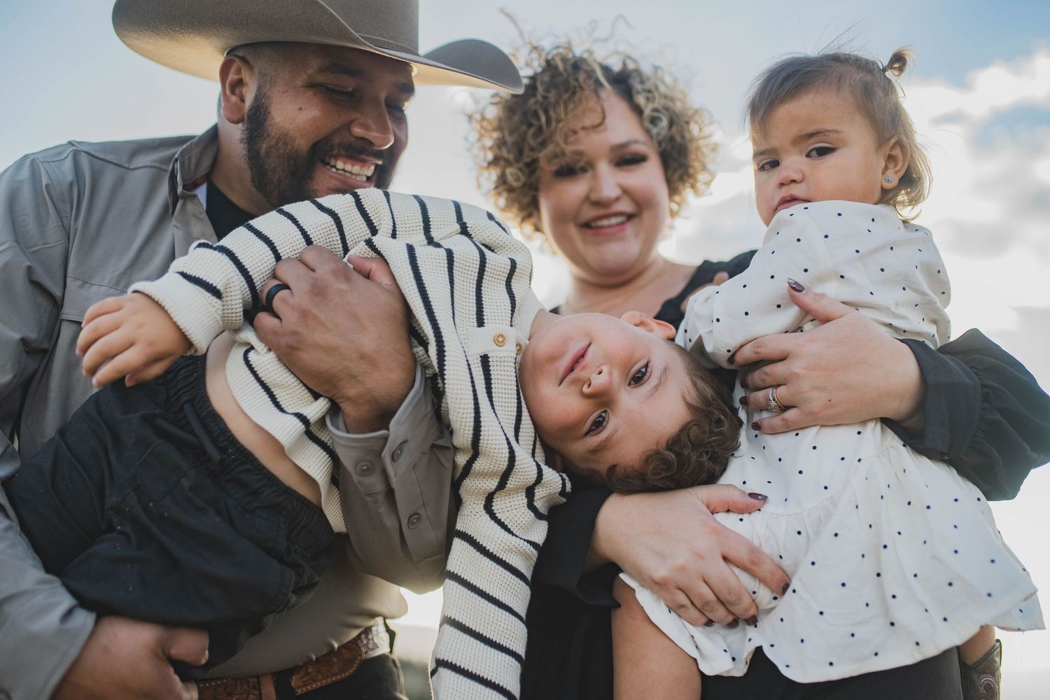 A cheerful family laughs and embraces together outdoors against a bright blue sky.
