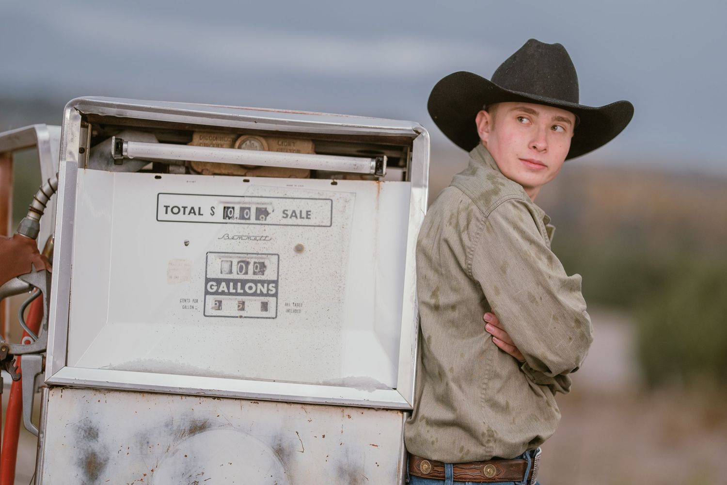 A cowboy in western attire stands against a rustic background with ranch equipment and storage containers.