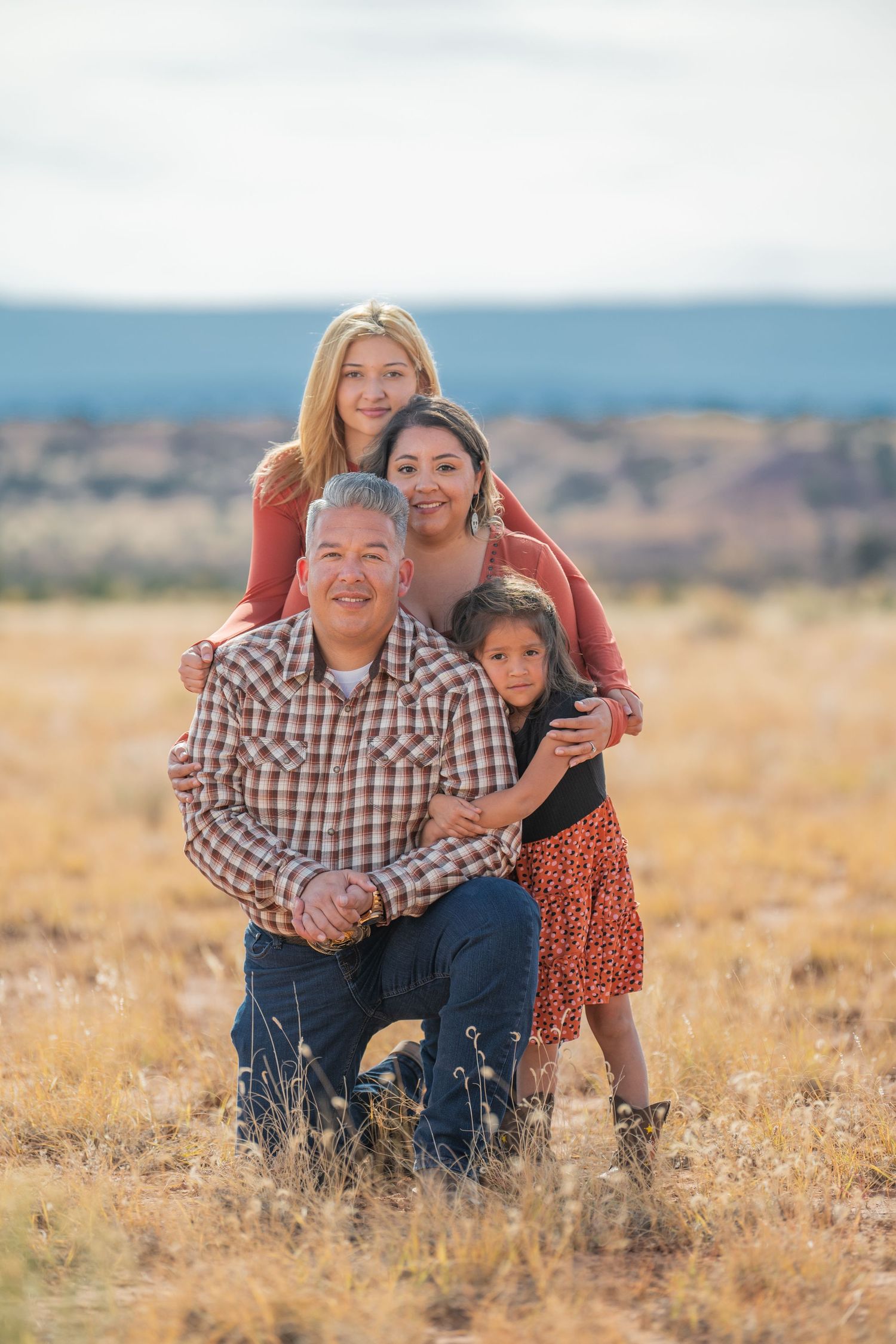 Family of three poses together in a golden autumn field during sunset, wearing coordinated casual outfits.