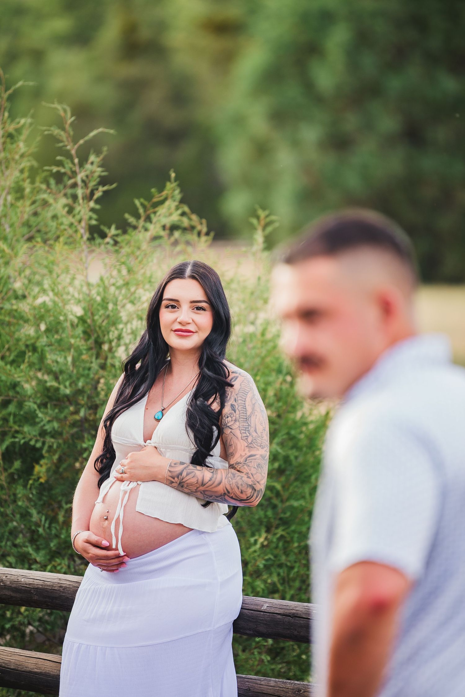 Pregnant woman in white dress with tattoos smiles outdoors near wooden fence, man in white shirt blurred in foreground.