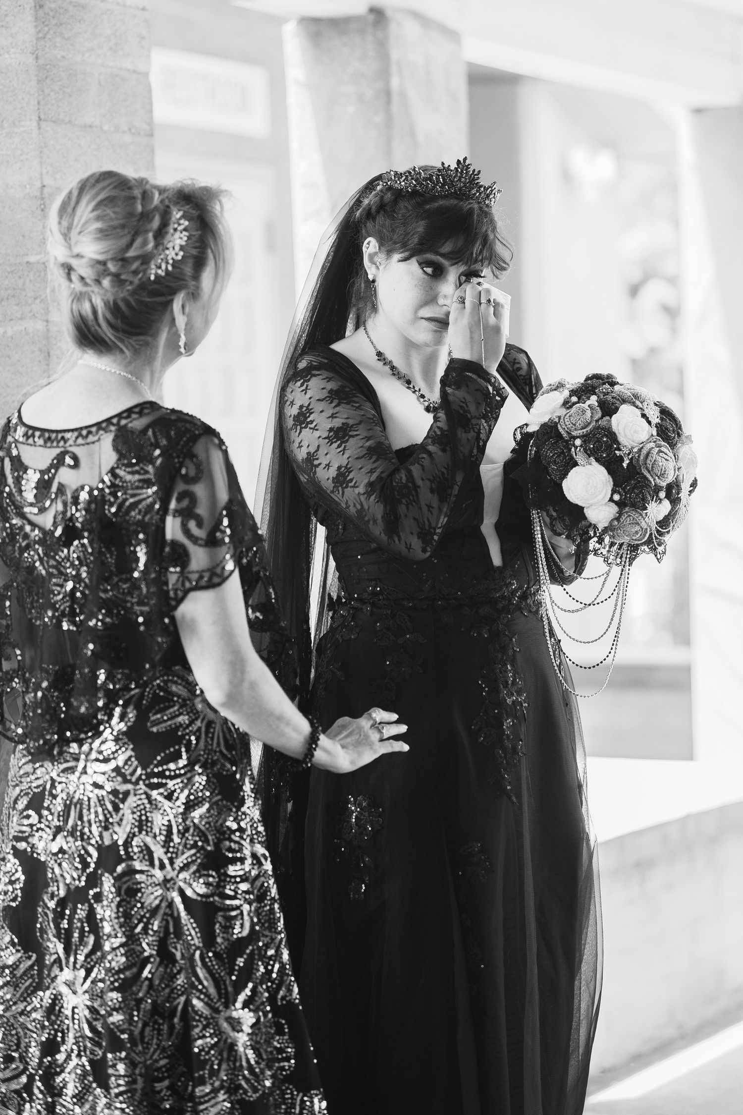 Black and white photo of a gothic bride in black lace gown holding bouquet, wiping tears beside woman in sequin dress.