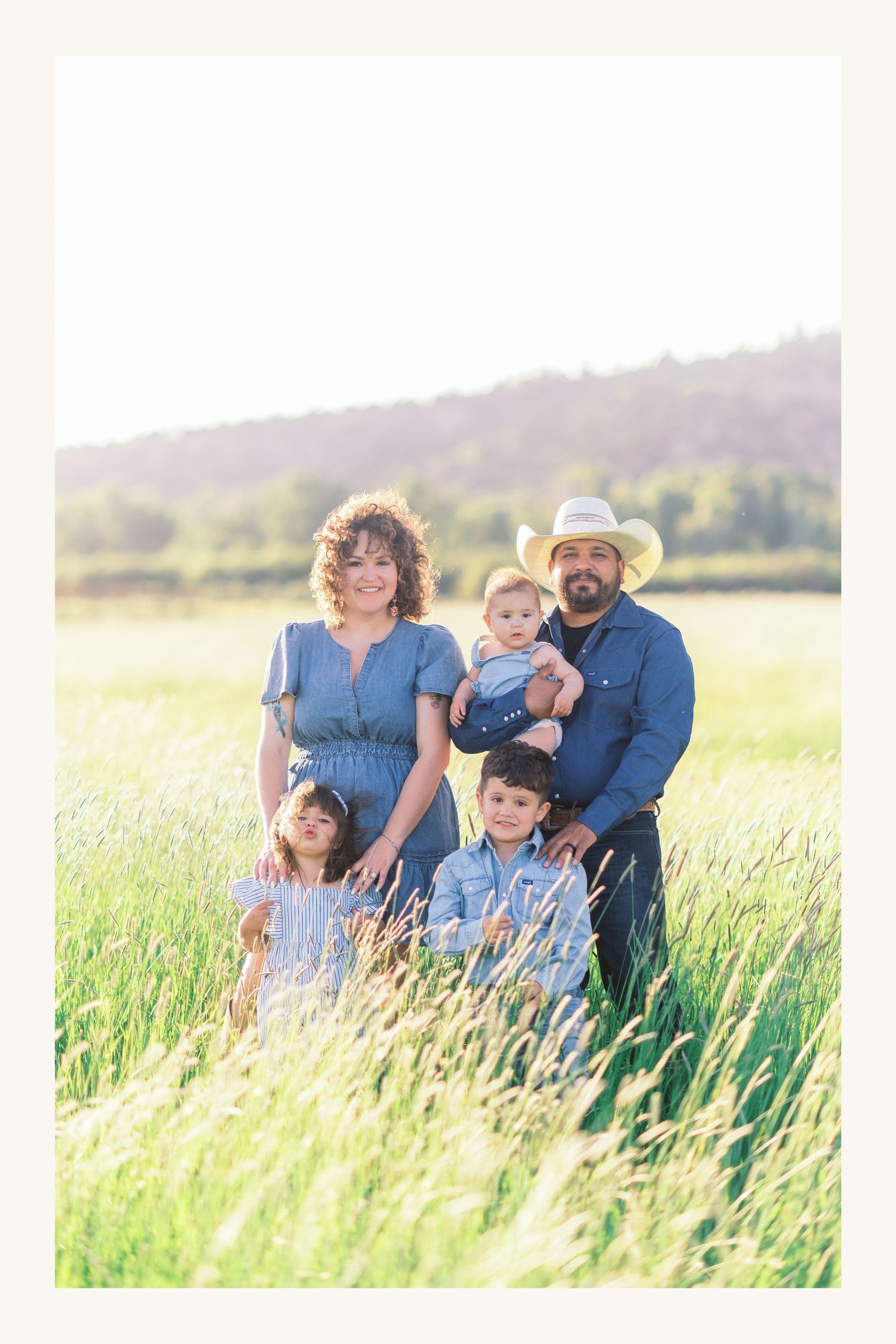 Family of five posing in a sunlit green field, parents in denim with kids, father wearing a cowboy hat.