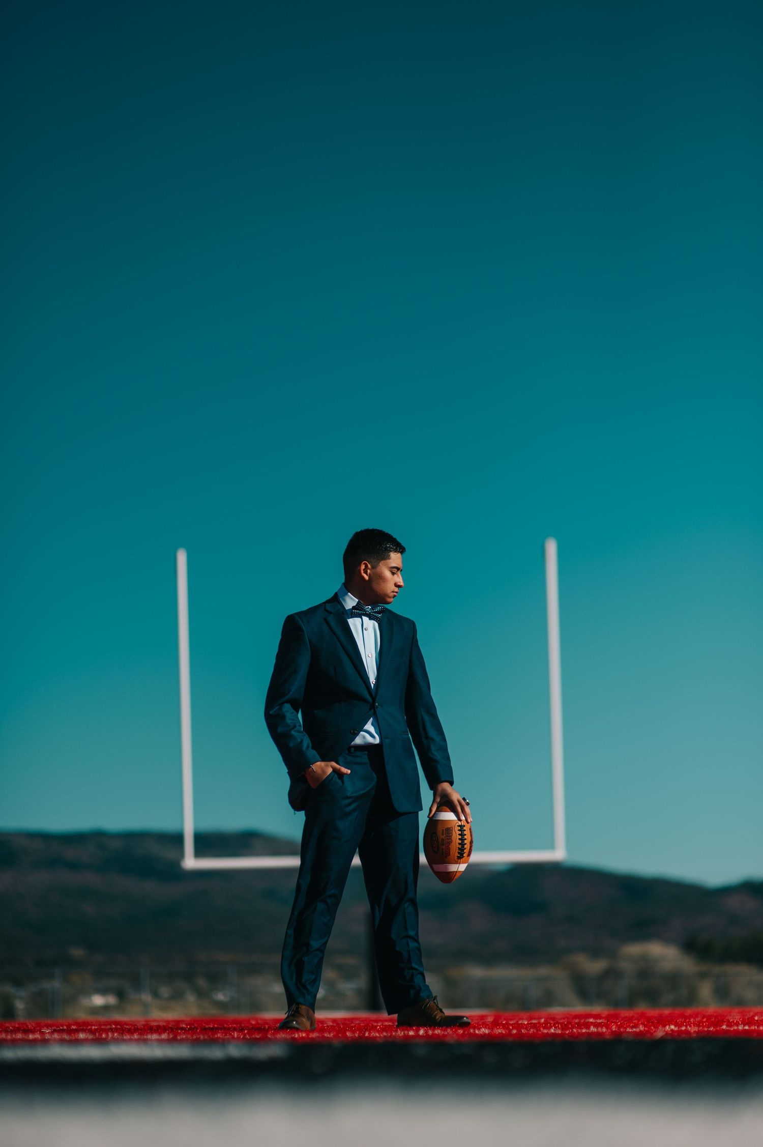 Man in dark suit holding football on field with goal posts and blue sky backdrop.