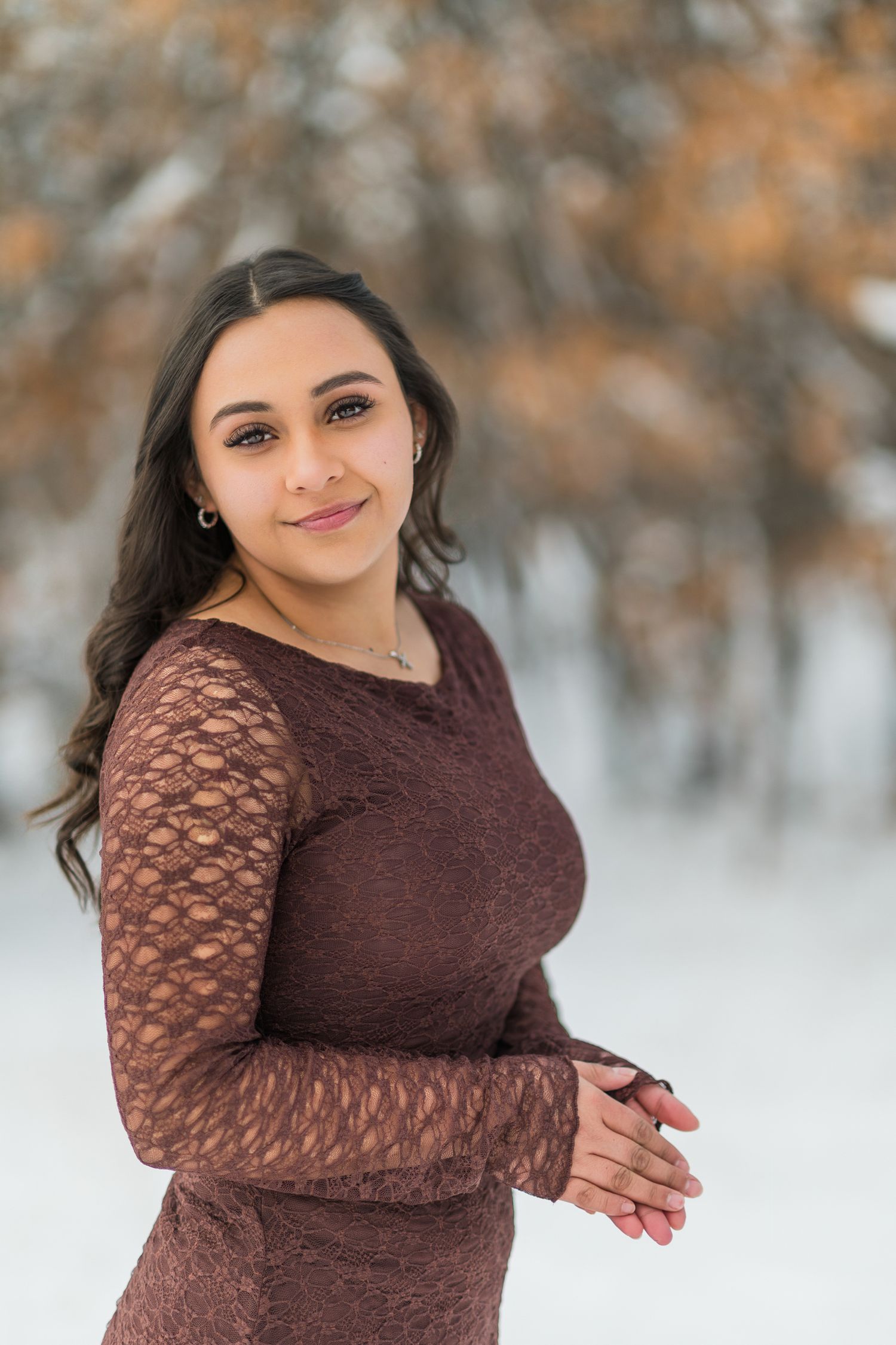 A woman in a brown lace dress poses outdoors in a snowy winter setting with a warm smile.