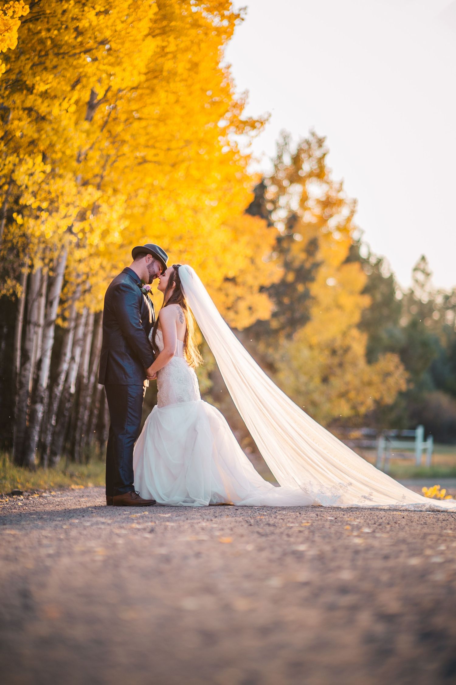 A romantic wedding portrait with a couple embracing on a dirt path lined with golden autumn trees.
