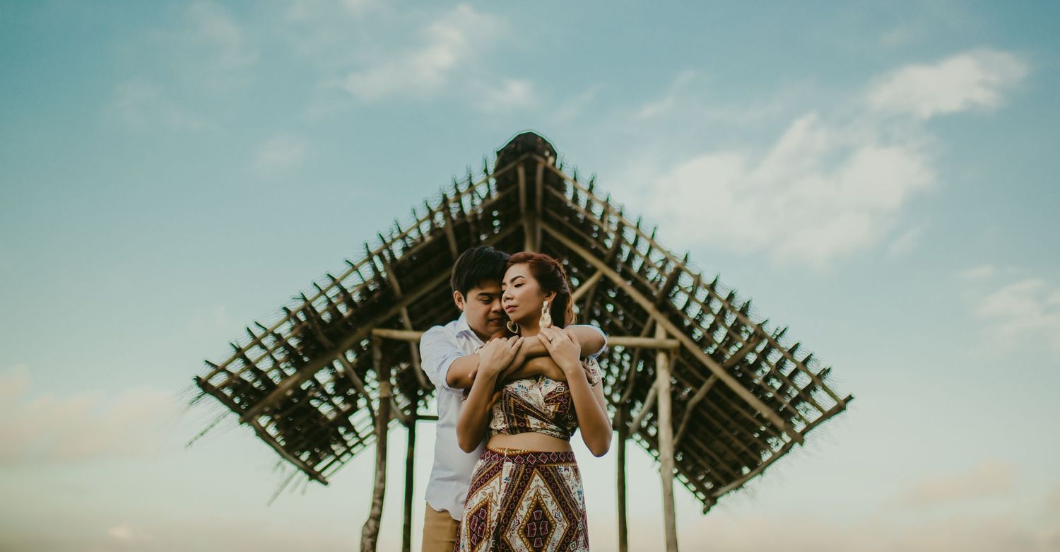 A couple embracing beneath a rustic wooden structure against a blue sky.