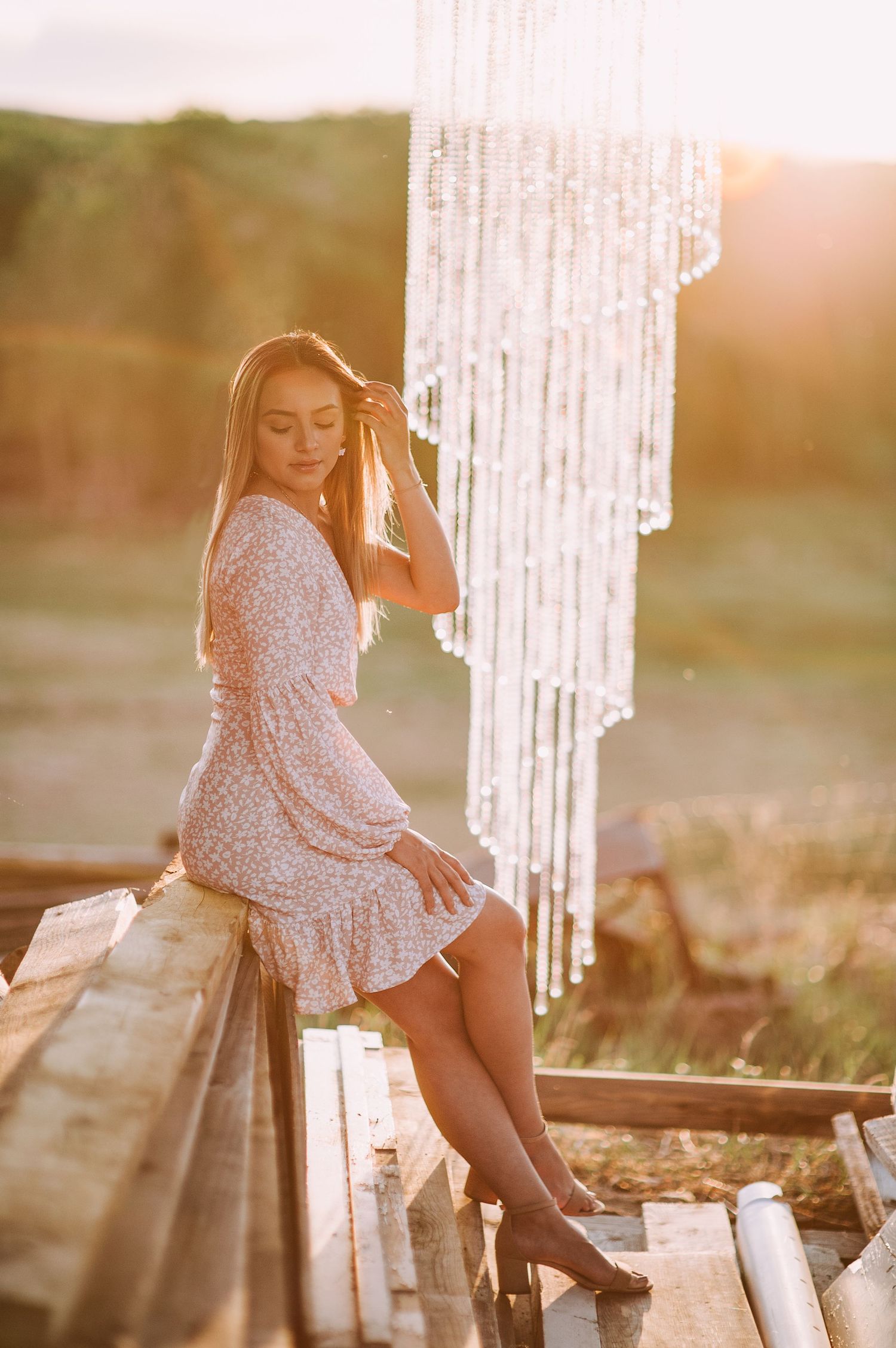Person in floral dress poses against hanging crystal backdrop during sunset.