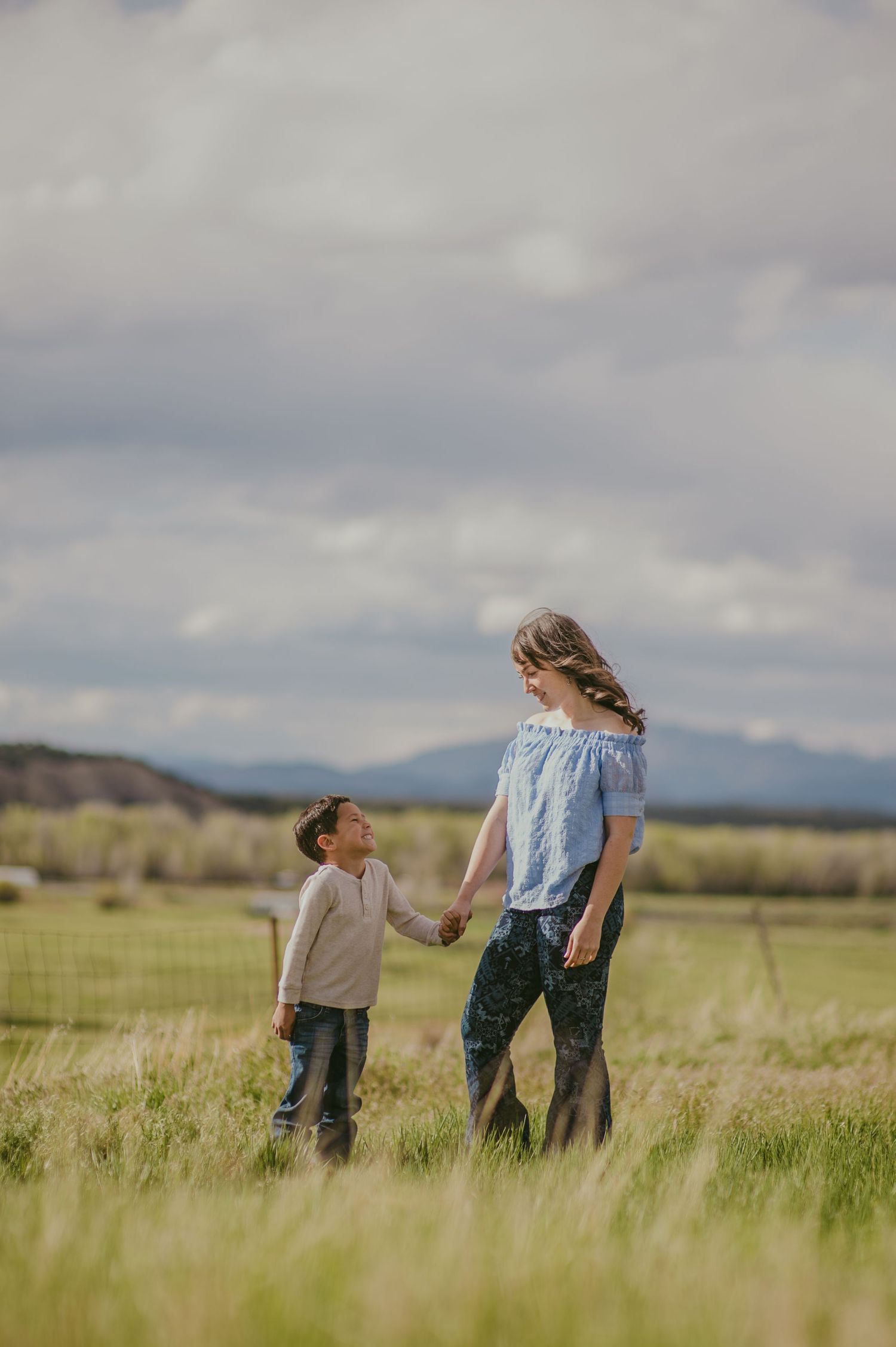 Mom holding her toddler son's hand as they stand in a green meadow in Los Ojos — natural light portrait by The Amoras.