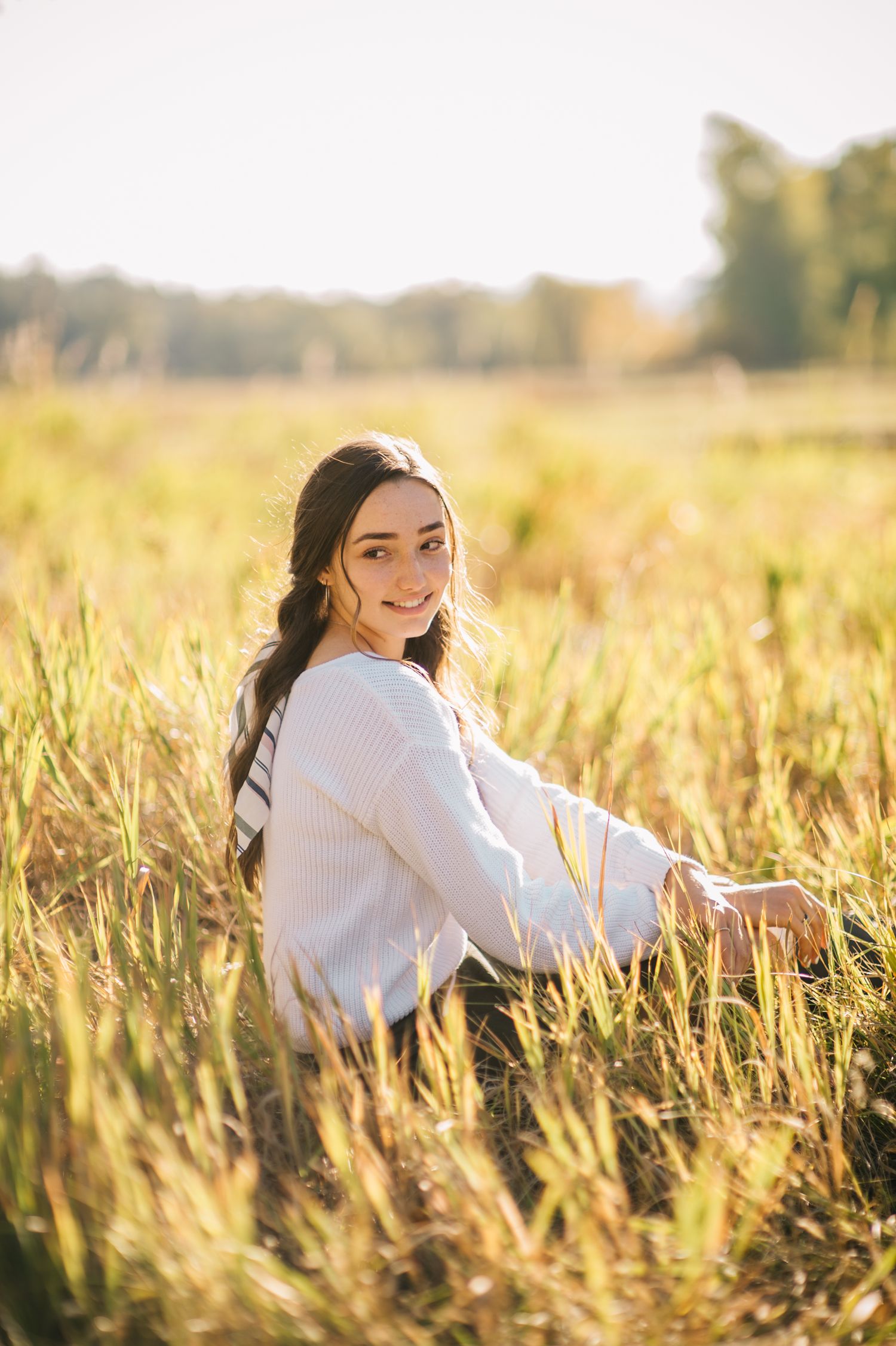 Senior girl sitting in a sunlit grass field, captured in soft natural light by Quiet Light by The Amoras.