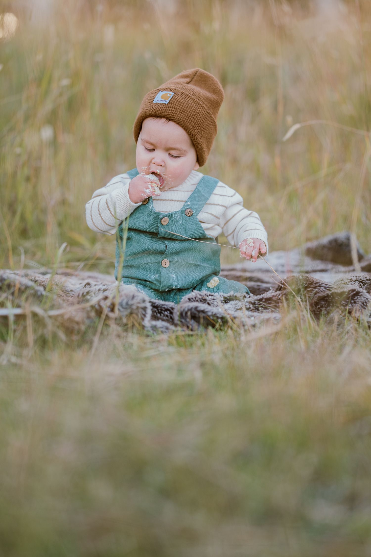 Small figure in green overalls and brown beanie lying on rustic wooden log in grassy outdoor setting.