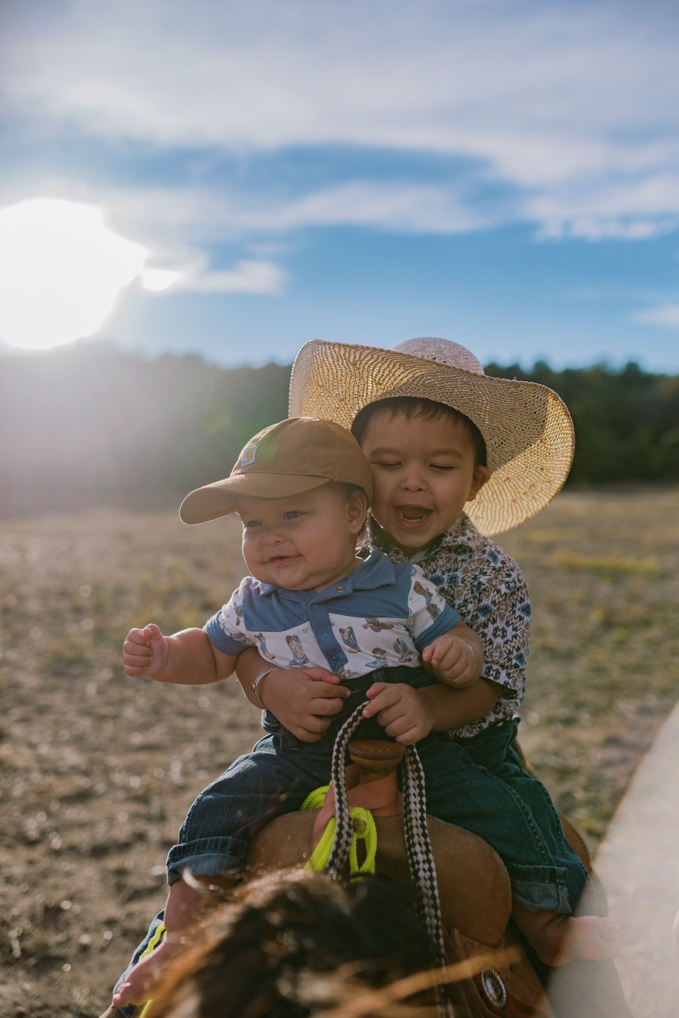 Two young toddlers in cowboy hats sitting together outdoors on a sunny day in a rural field.