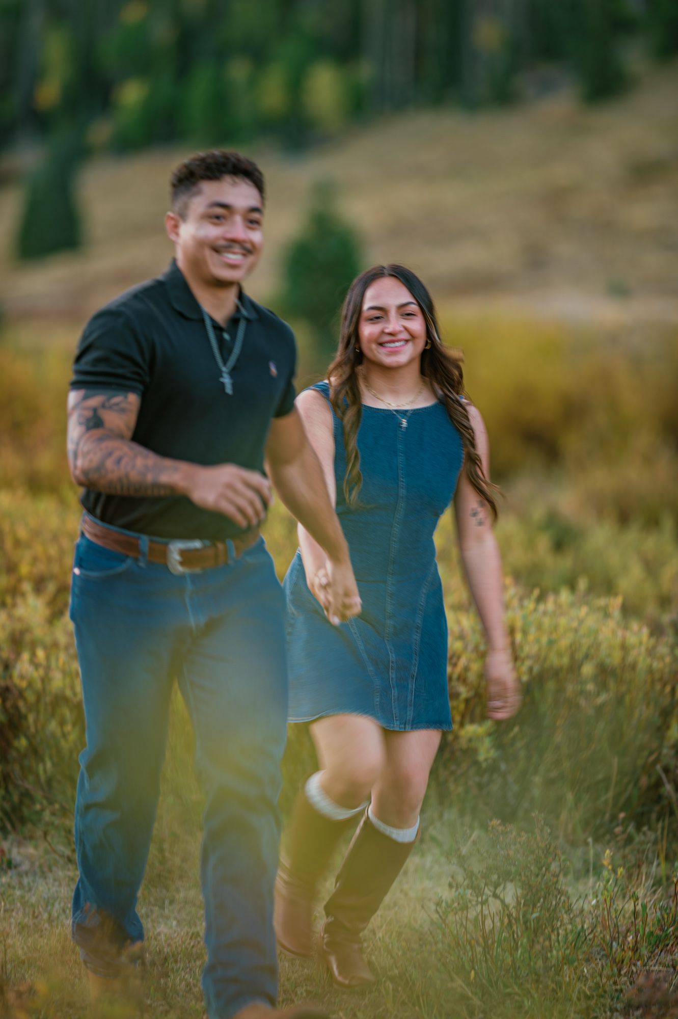A smiling couple holds hands while walking through a scenic mountain meadow, the woman wearing a denim dress and tall boots.