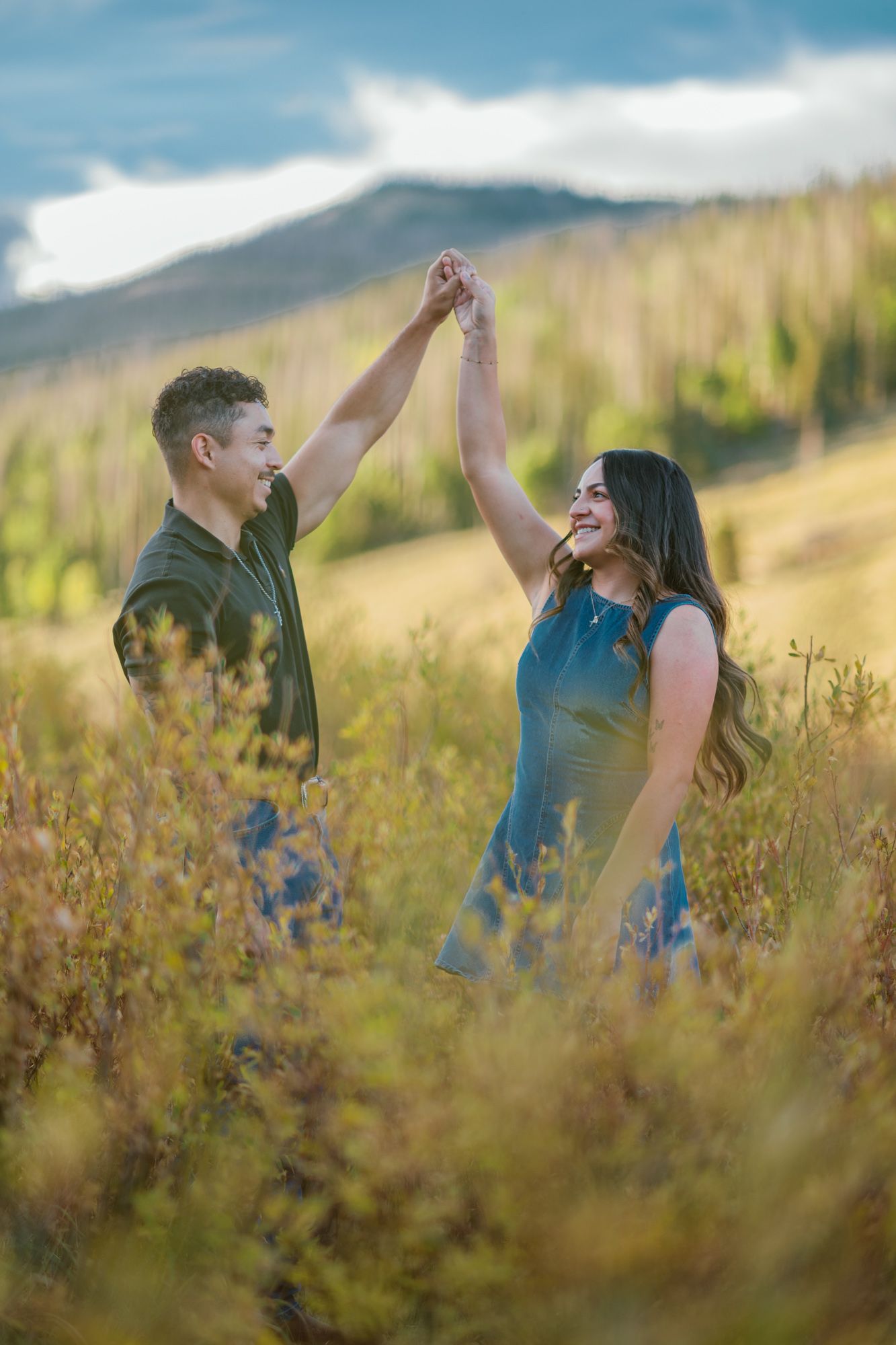 Couple dancing outdoors in a mountain meadow, man spinning woman with raised hands, surrounded by golden wildflowers.