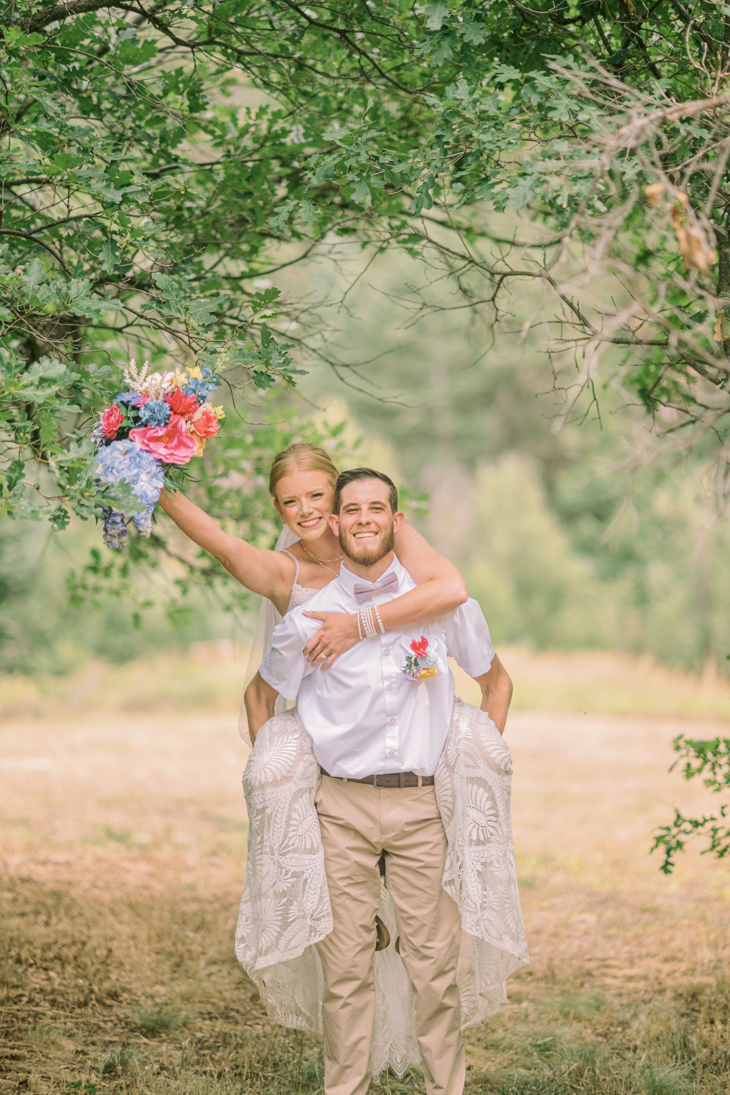 Groom giving bride a piggyback ride outdoors, bride holding colorful bouquet, both smiling, surrounded by green trees.