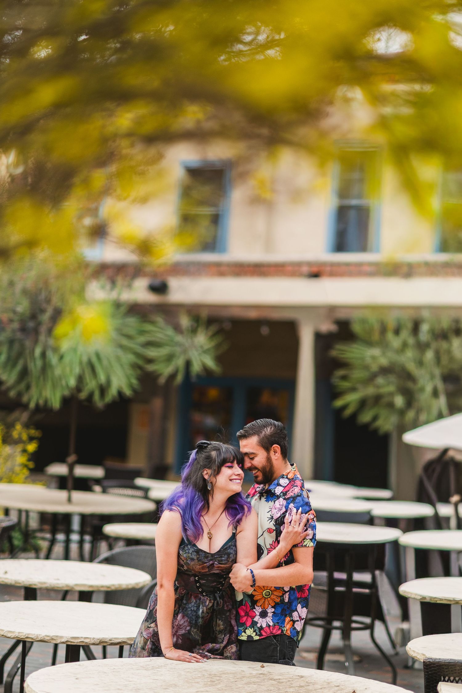 Couple embracing and smiling at an outdoor cafe patio, woman with purple hair in floral dress, man in colorful shirt.