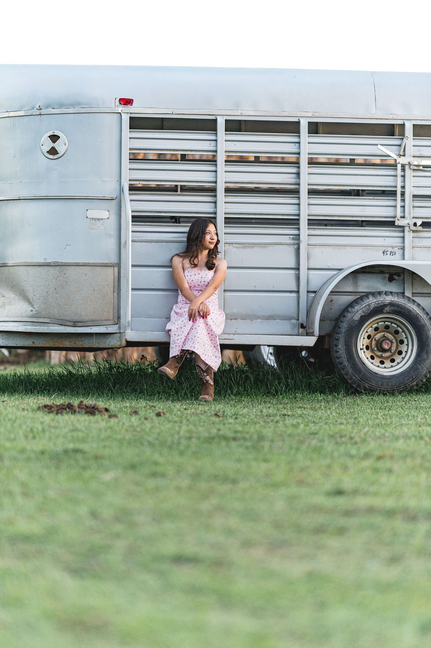 A person sits on grass beside a silver livestock trailer, creating a rustic country scene outdoors.