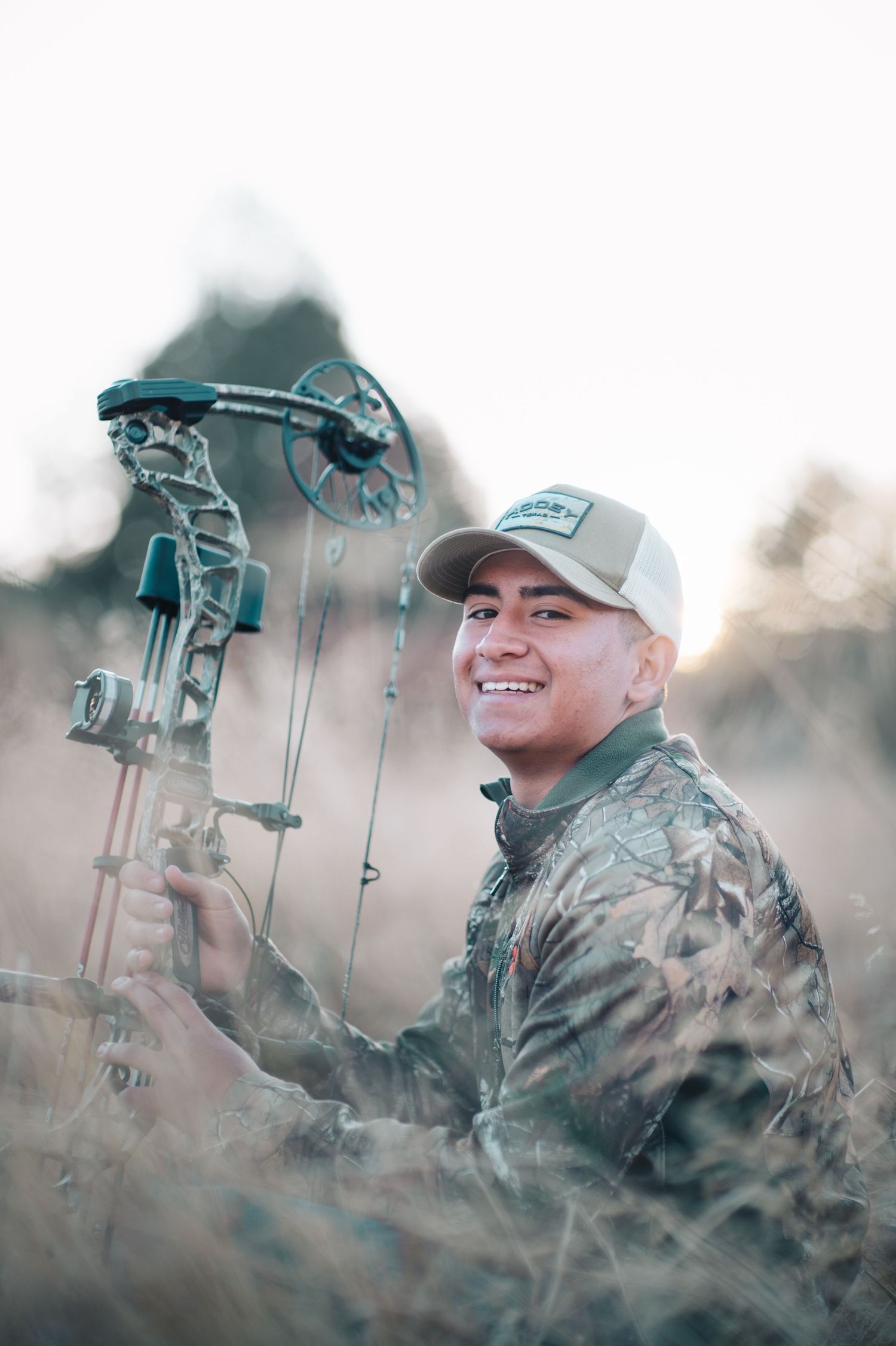Smiling hunter in camouflage holding a compound bow in a field at dusk.
