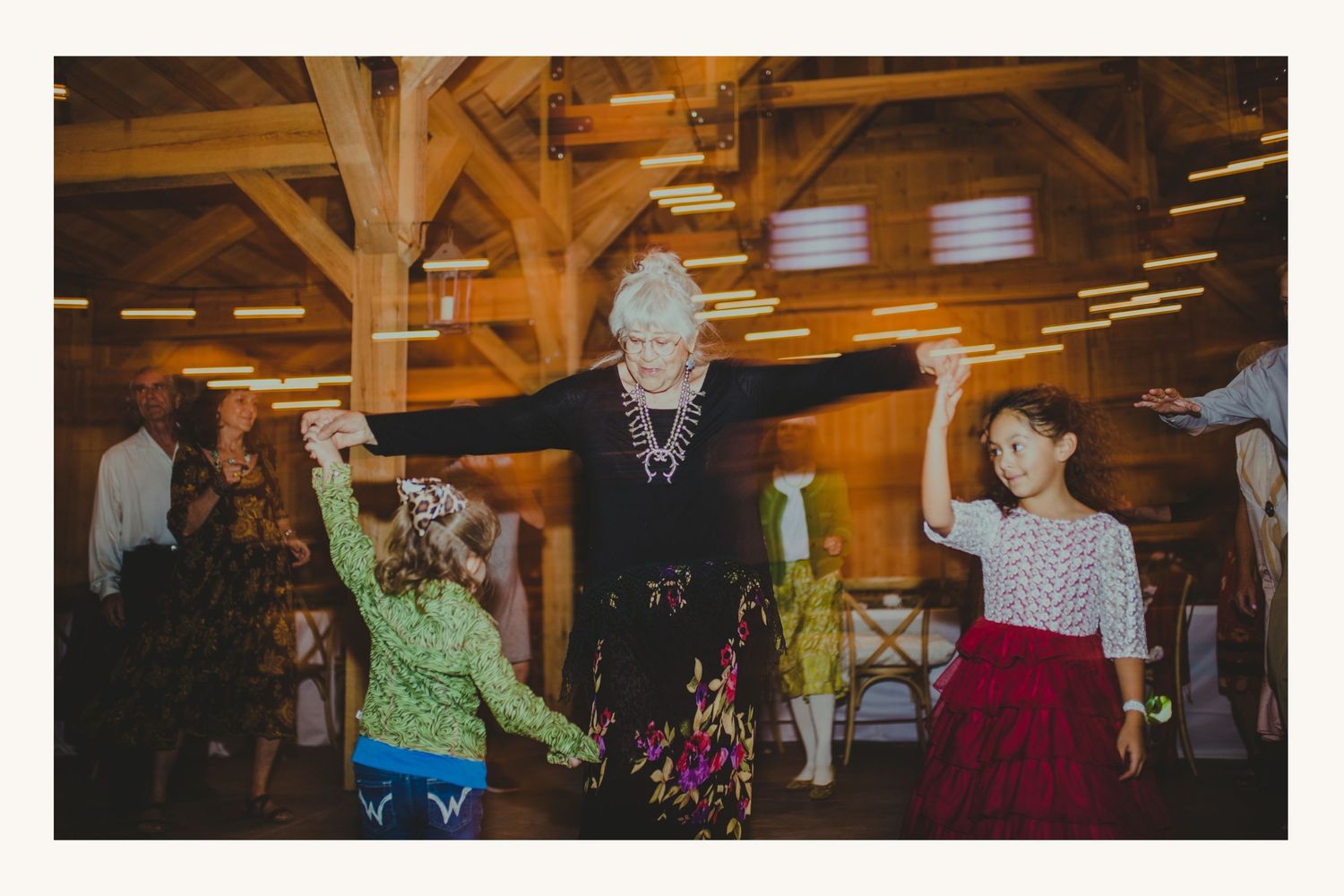 Elderly woman joyfully dancing with two young girls in a rustic barn wedding reception venue.