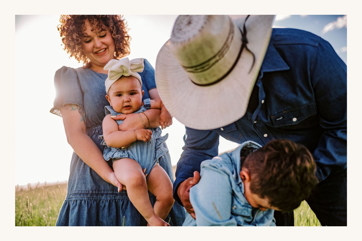 Family outdoor photo with mother holding baby girl, father in cowboy hat, and young son in green field.