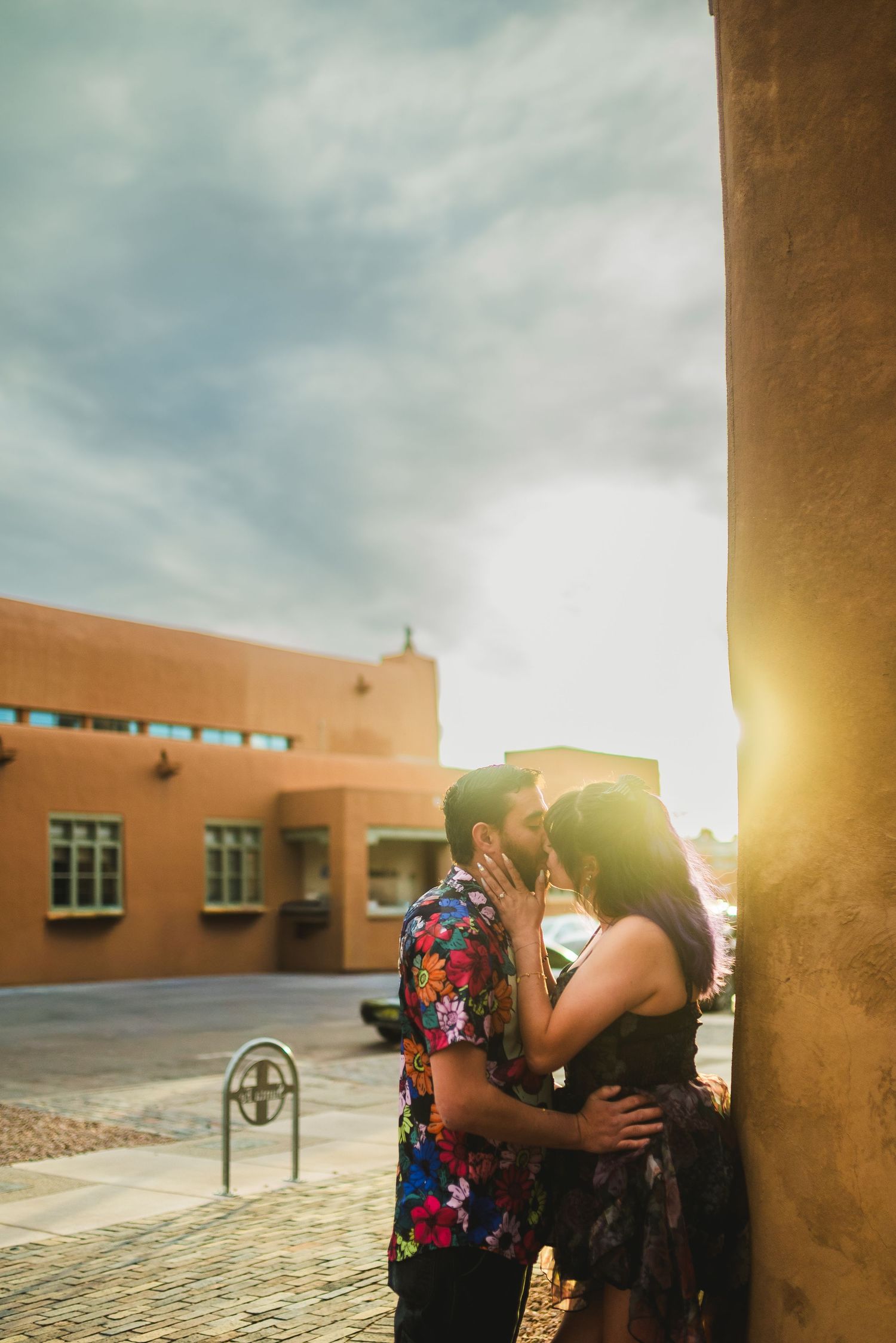 Couple sharing a romantic kiss against a golden sunlit adobe wall during a warm sunset engagement photo session.