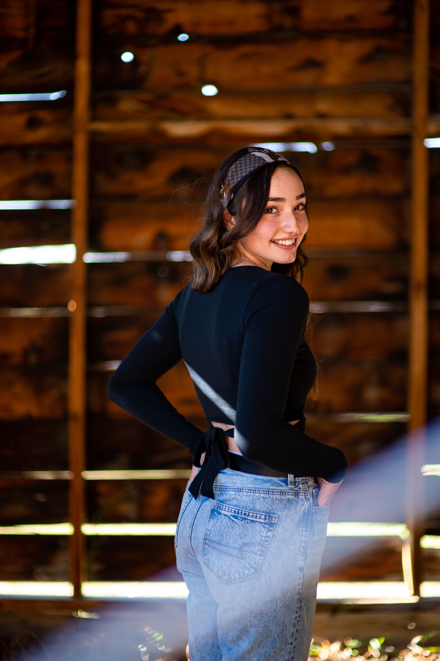 Person in dark top and jeans poses in a rustic barn with wooden beams and natural lighting.
