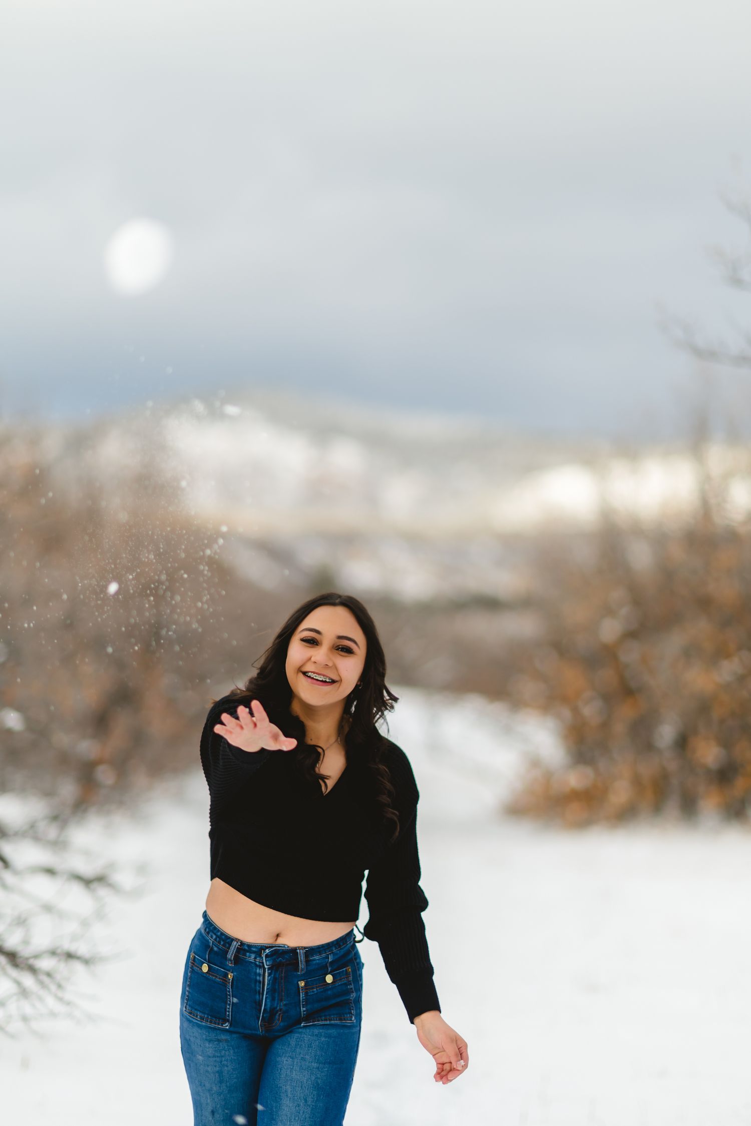 Person in black sweater and blue jeans plays joyfully in winter snow landscape with blurred background.