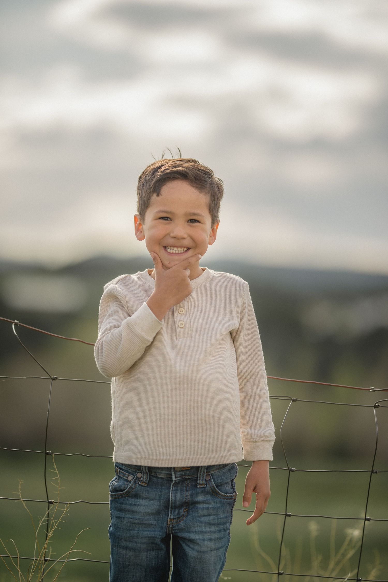 Young child in cream sweater and jeans smiles while standing by a wire fence in an outdoor rural setting at dusk.