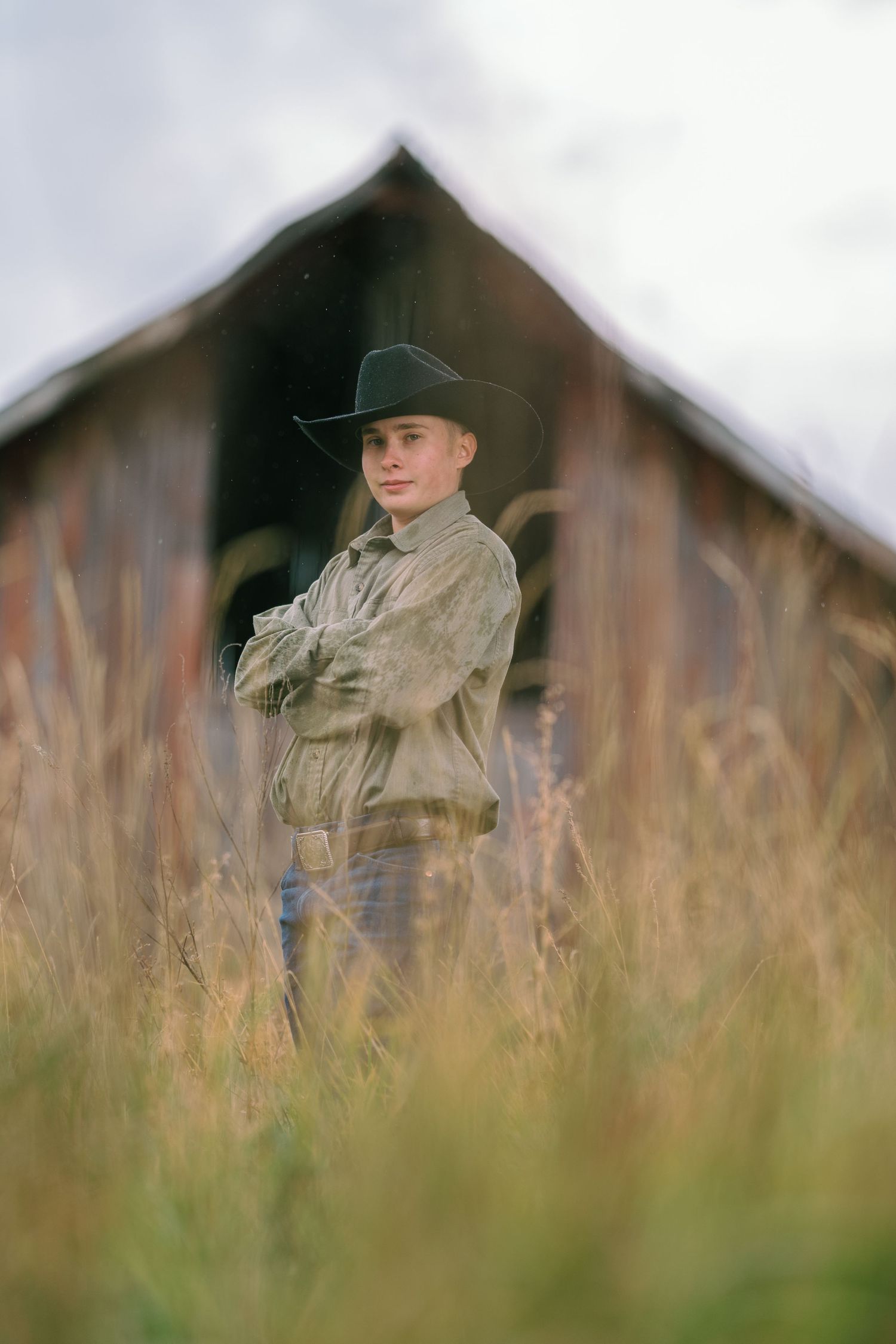 Person in cowboy hat and outdoor clothing stands in tall grass with a rustic barn in the background.