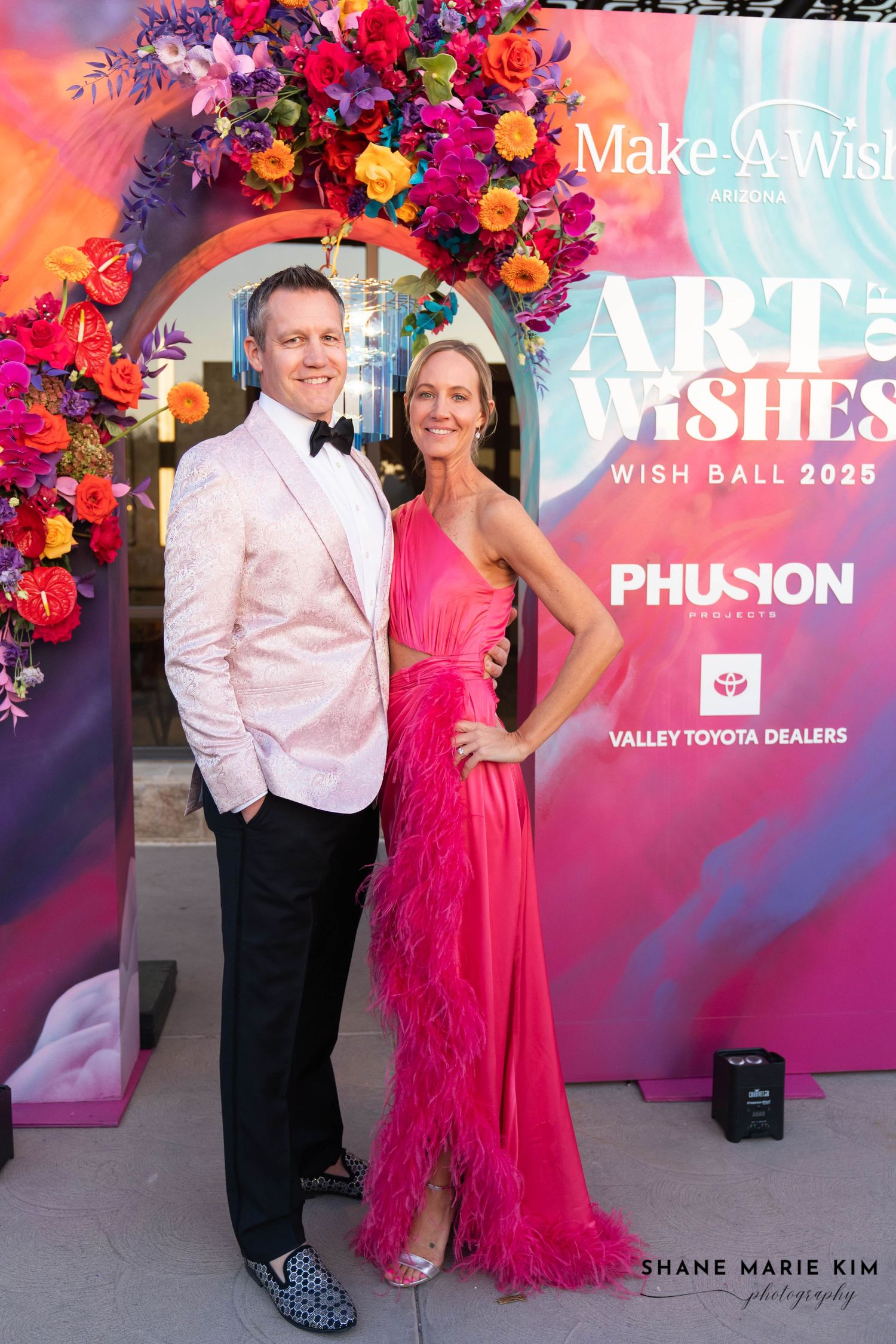 make a wish ball step and repeat photography, guy wearing a light pink blazer with black tie and pants, woman wearing a bright pink dress