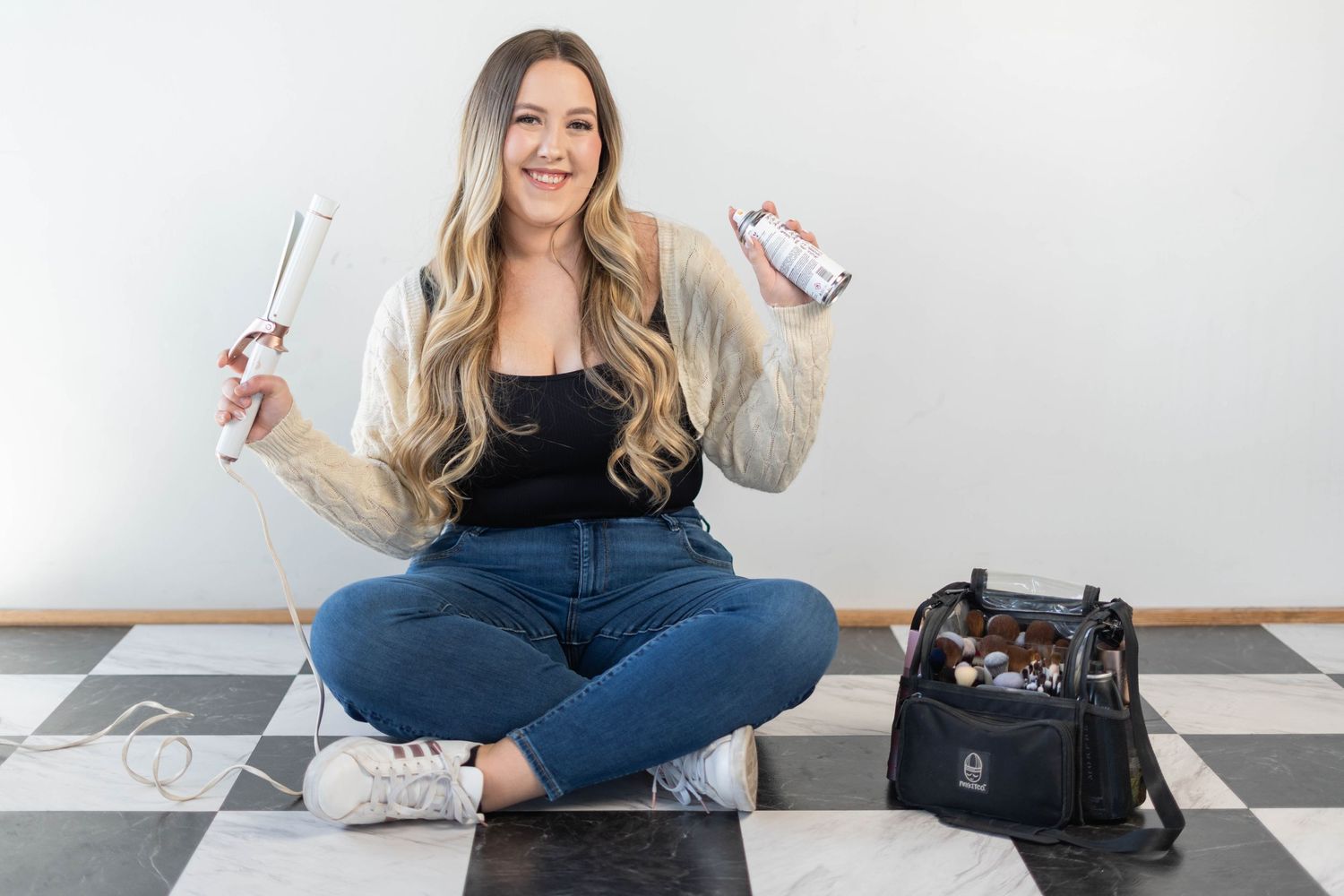 girl holding a hair styling tool and hair spray, make up back with make up brushes and make up tools, girl sitting on checkered floor