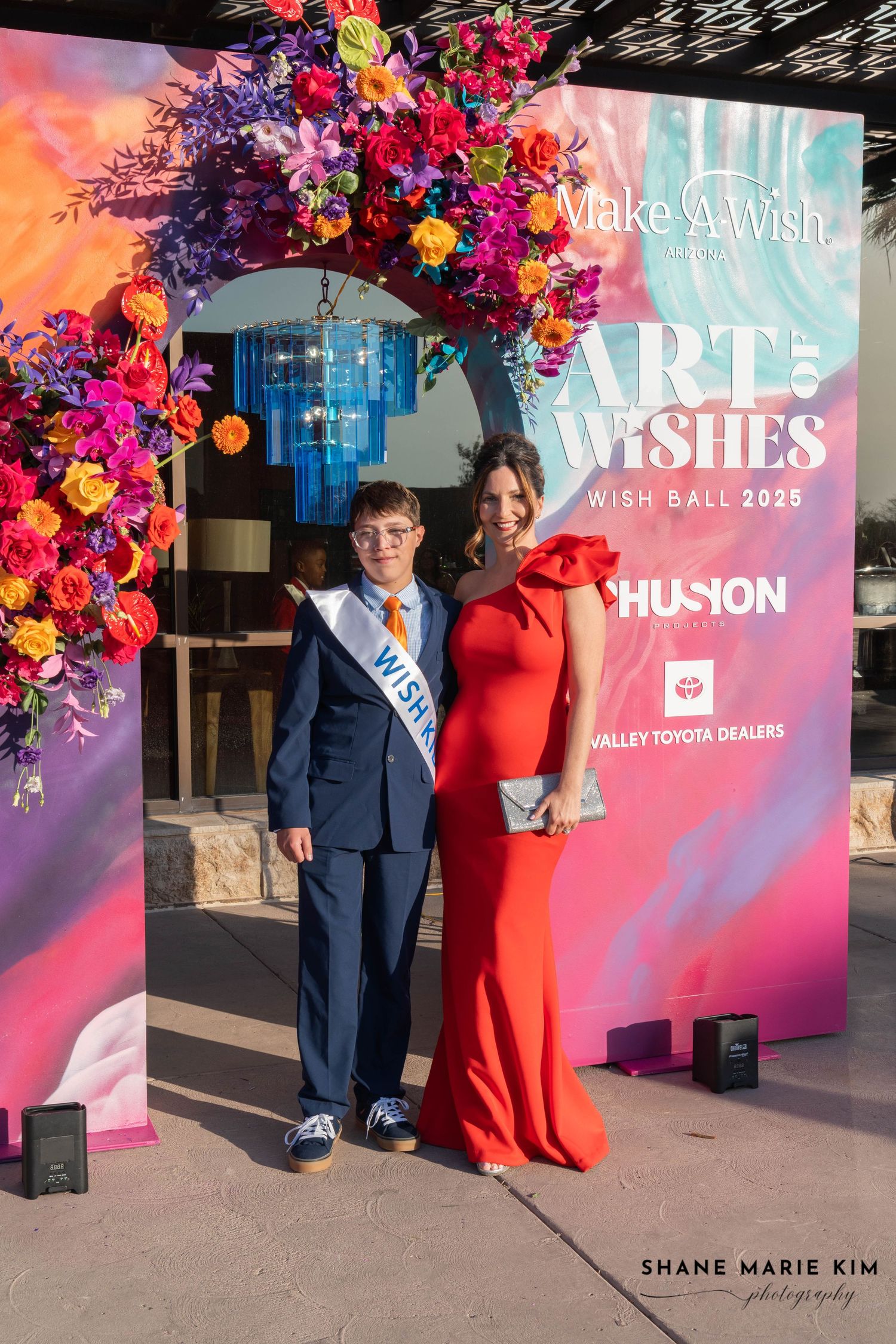boy with glasses wearing a blue pant suit with a red tie, woman wearing a red, one strap dress