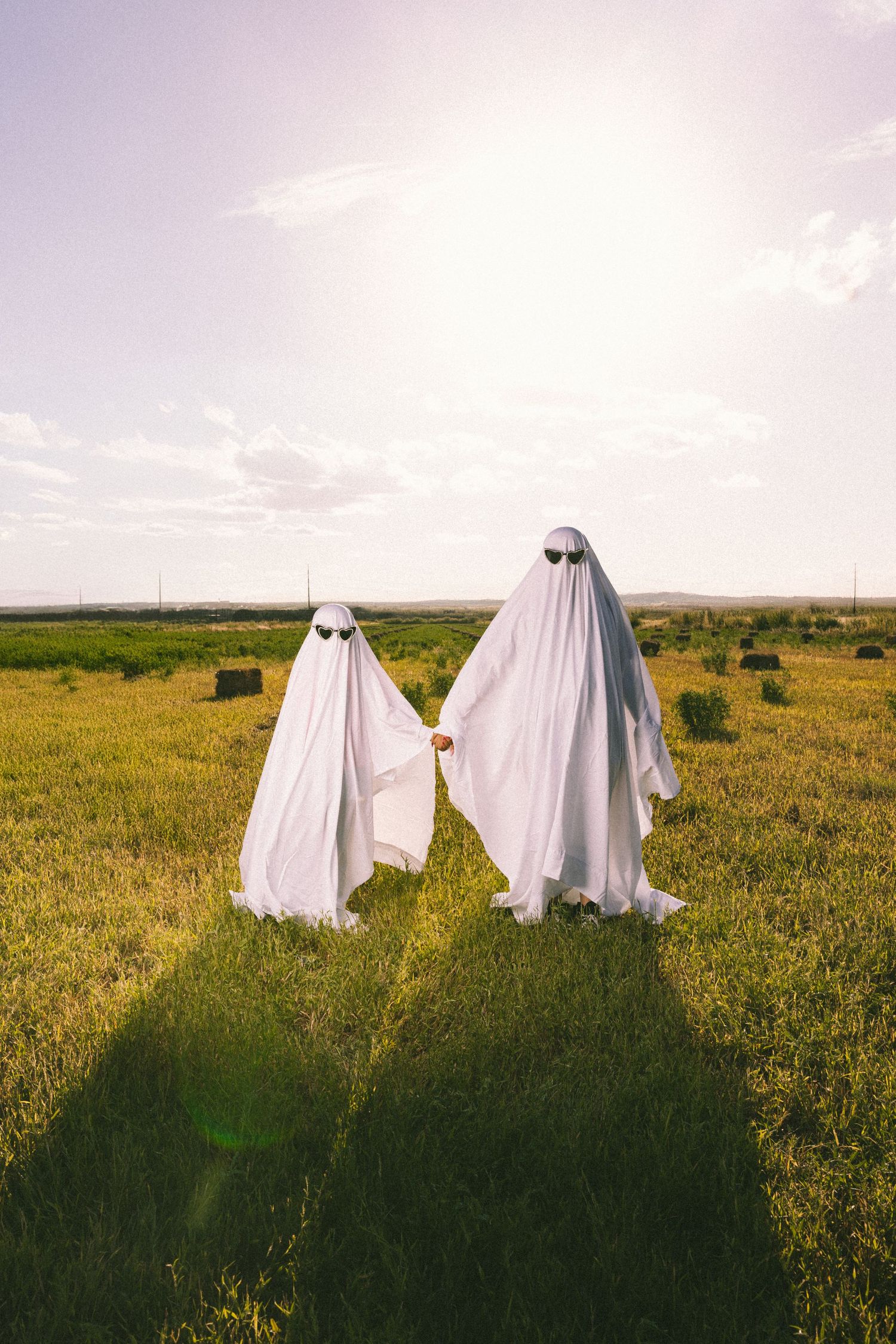 Ghost figures wearing white sheets pose in a sunny field with sunglasses.