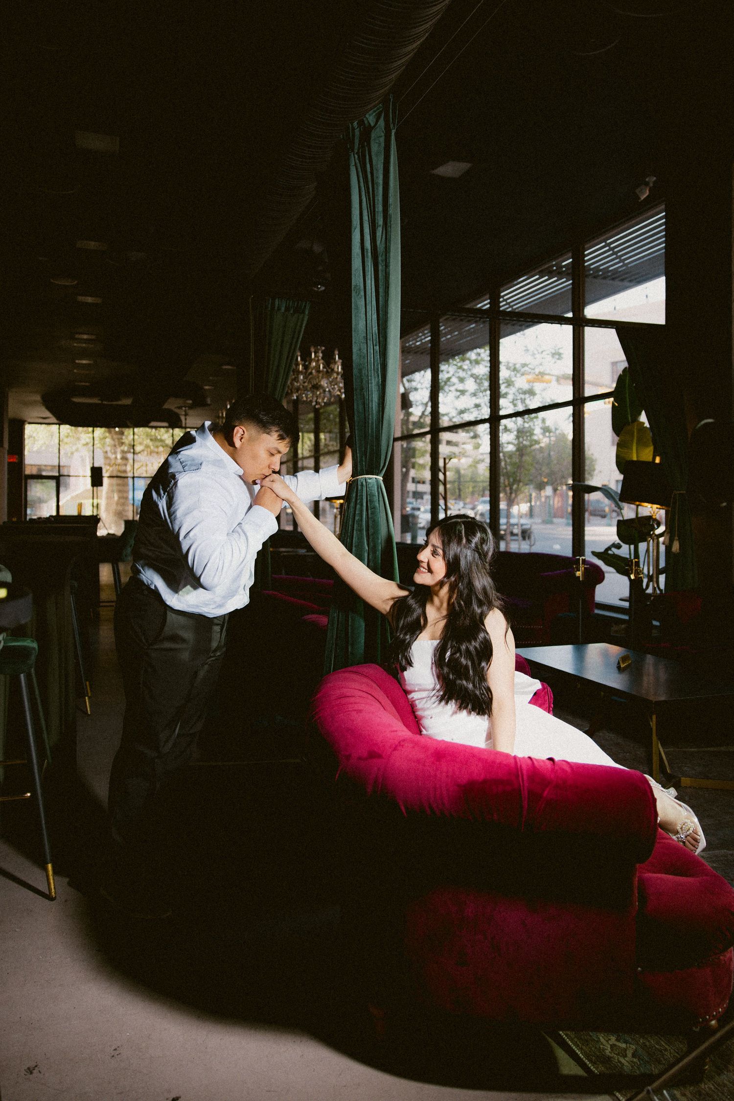 Couple in formal attire share romantic poses on a red velvet couch in a dimly lit venue.