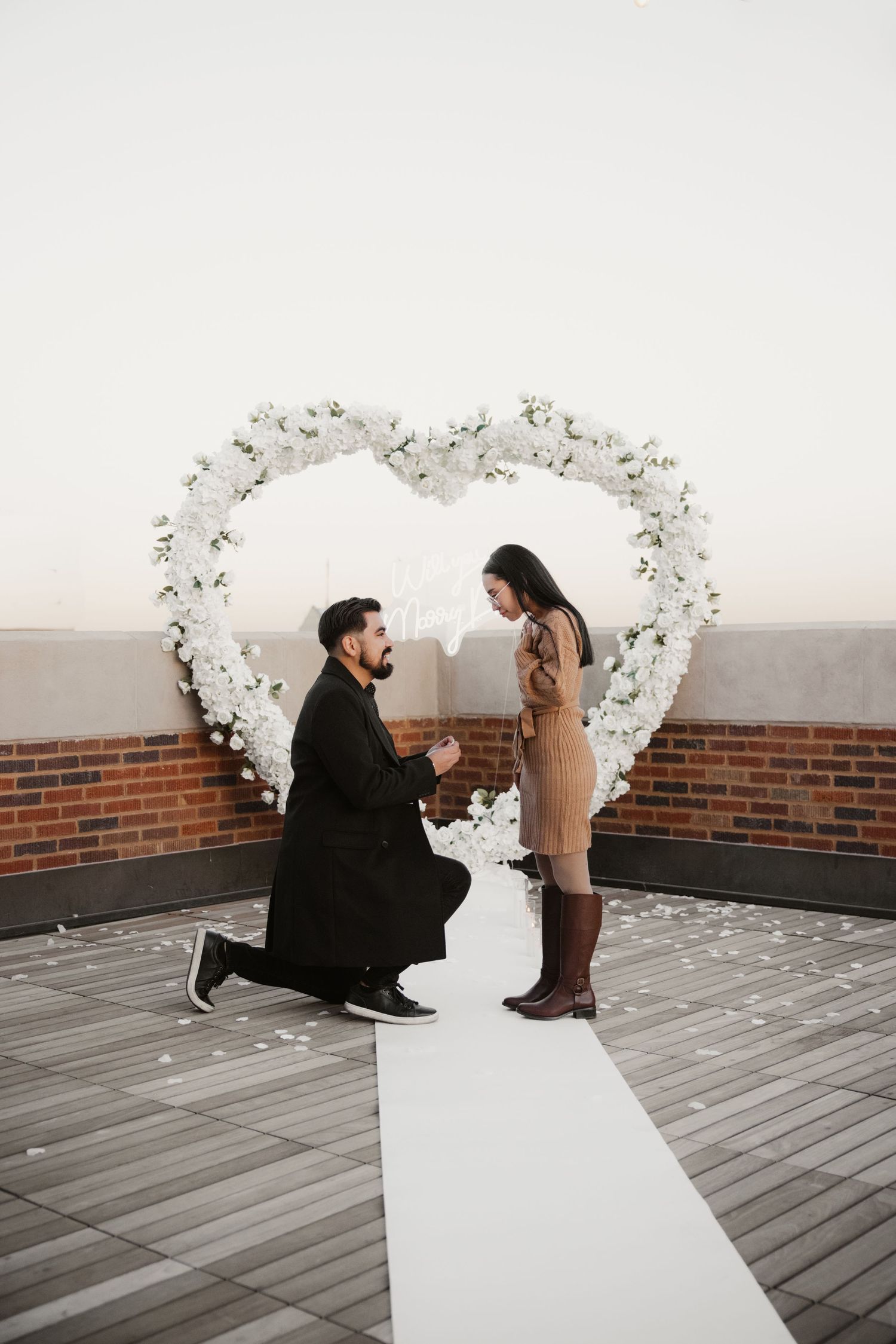 A romantic proposal scene under a white floral heart arch on a rooftop deck.
