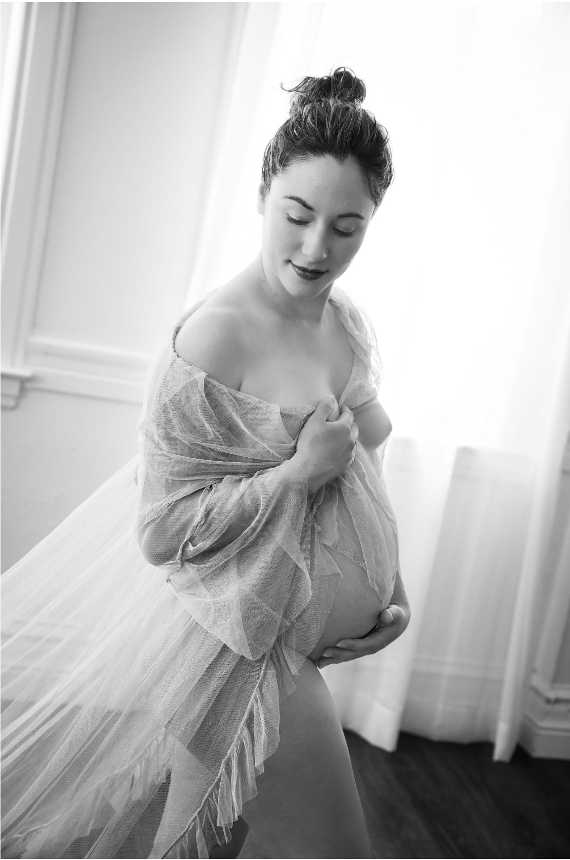 A black and white maternity photo showing an expecting mother in a flowing sheer dress near a window.