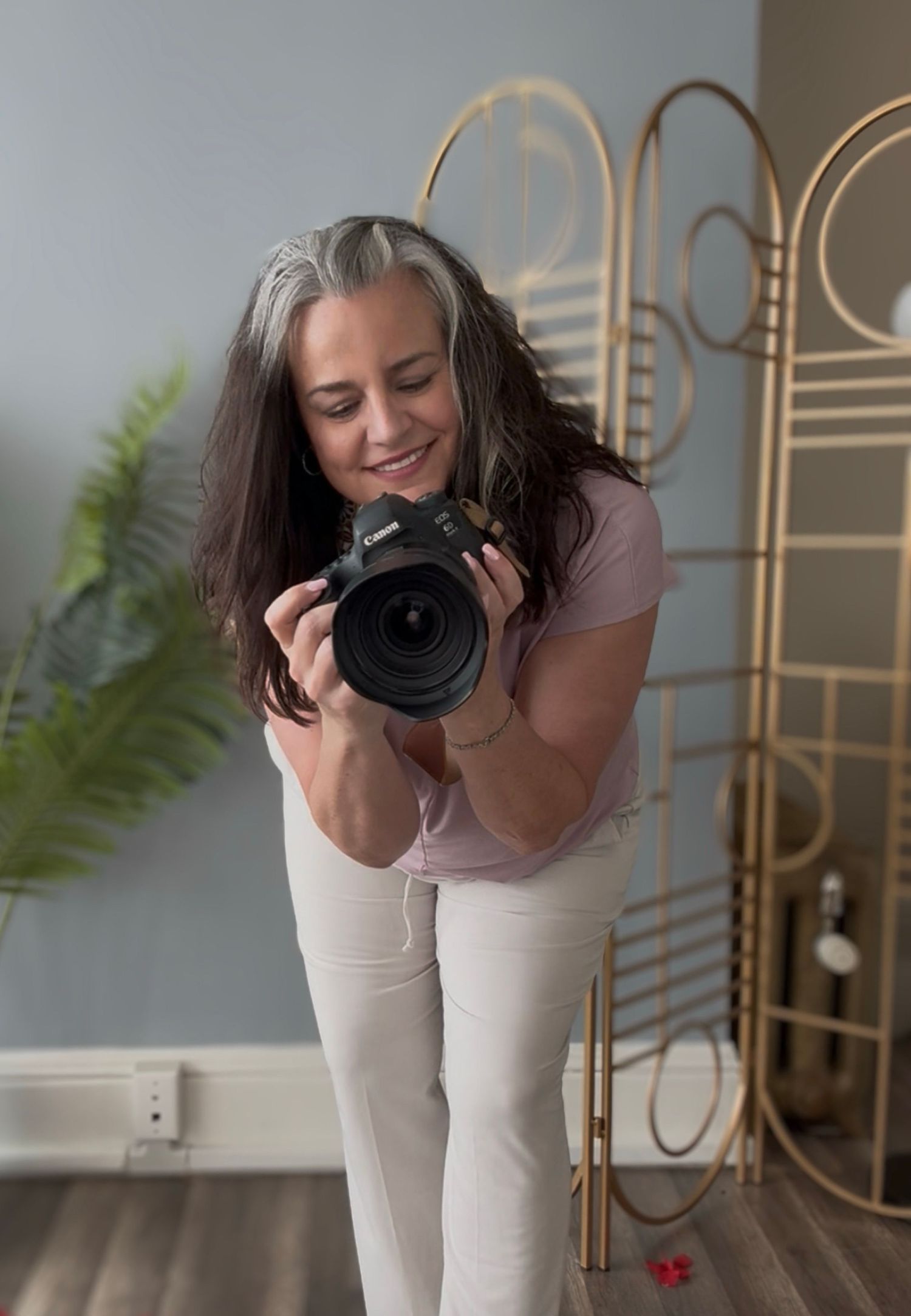 Photographer in casual attire smiles while holding a professional DSLR camera in a studio with gold room divider and plant decor.