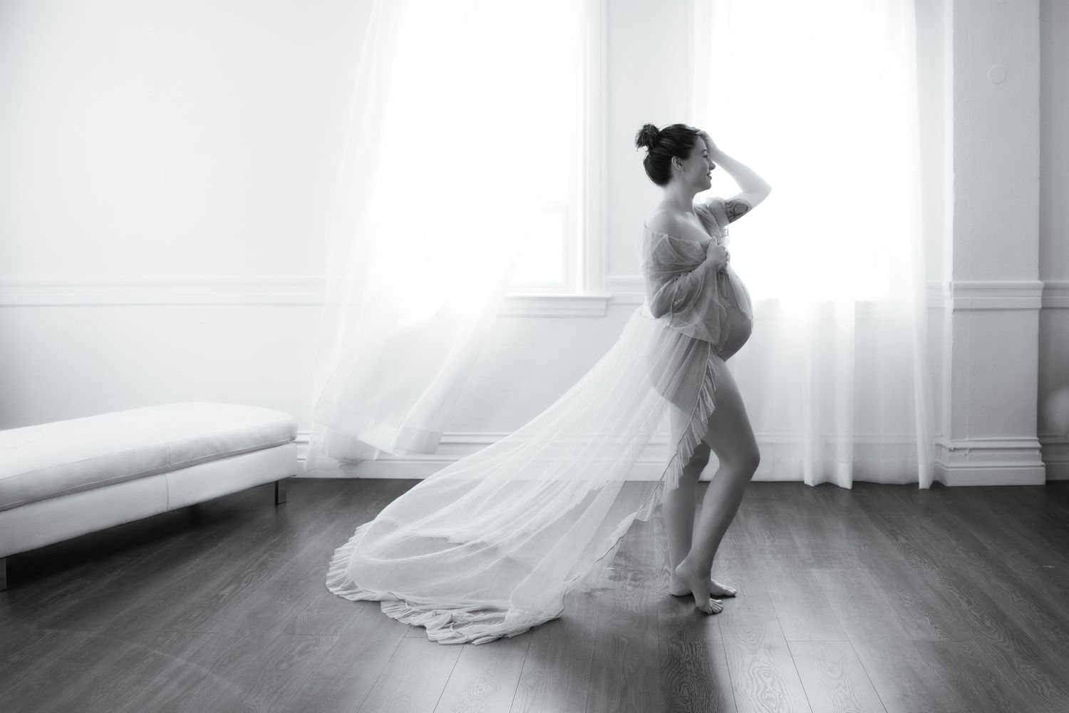 A graceful black and white portrait of an expectant mother in a flowing maternity gown standing by a window.