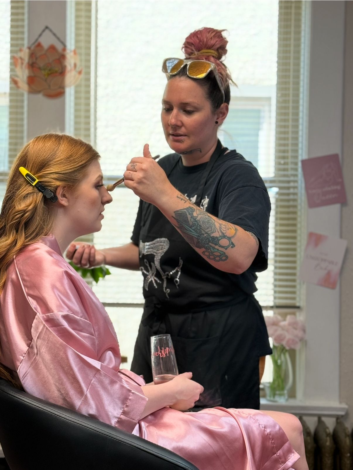 A hairstylist in black works on styling a client wearing a pink satin robe in a salon setting.