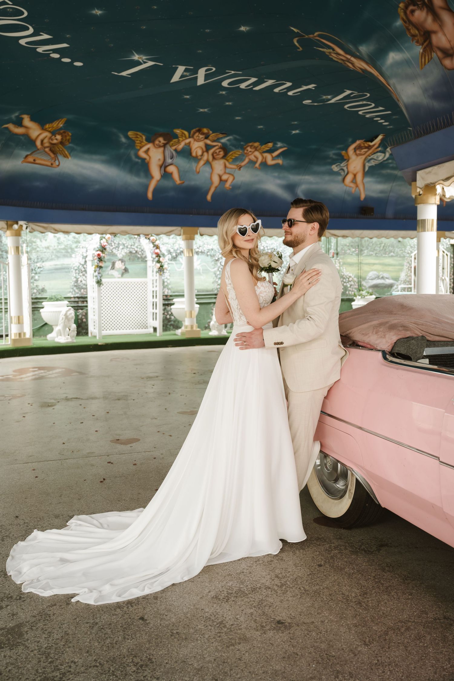 Stylish wedding couple poses with a pink vintage car under a decorated ceiling.