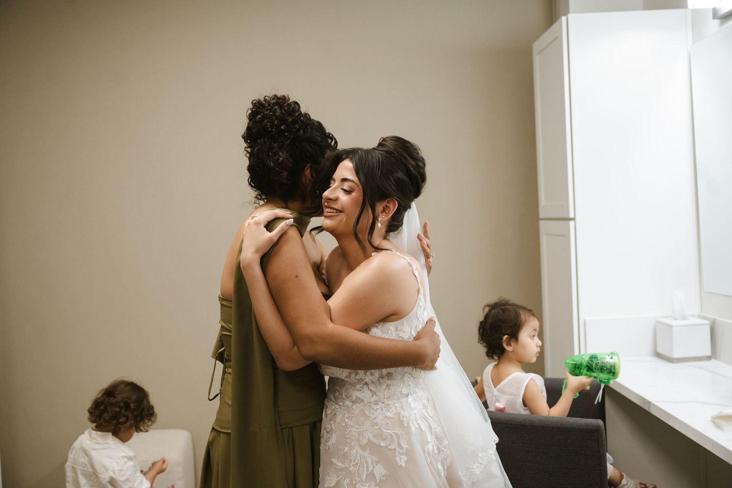 Bride in white lace gown shares an emotional hug with bridesmaid in olive dress in a bridal suite.