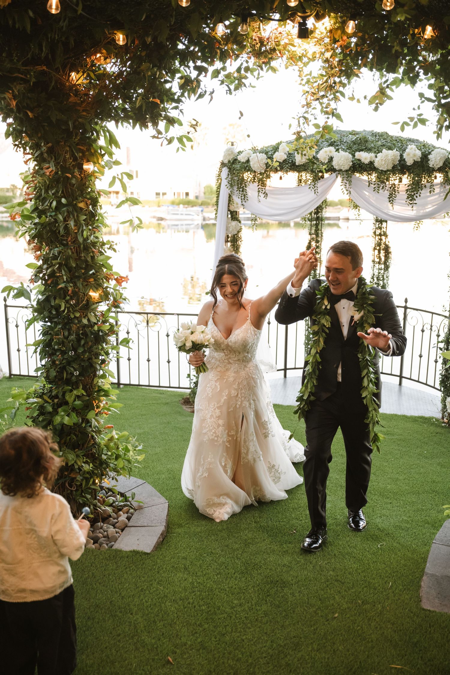 Bride and groom celebrate under floral arch by waterfront, raising hands joyfully after outdoor wedding ceremony.