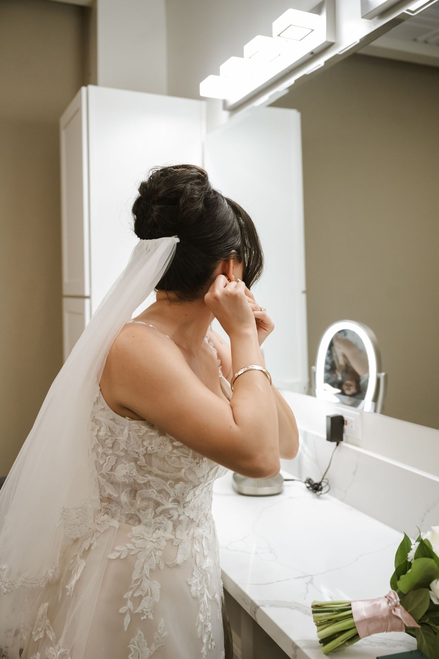 Bride in lace wedding gown and veil putting on earrings at vanity mirror before wedding ceremony.