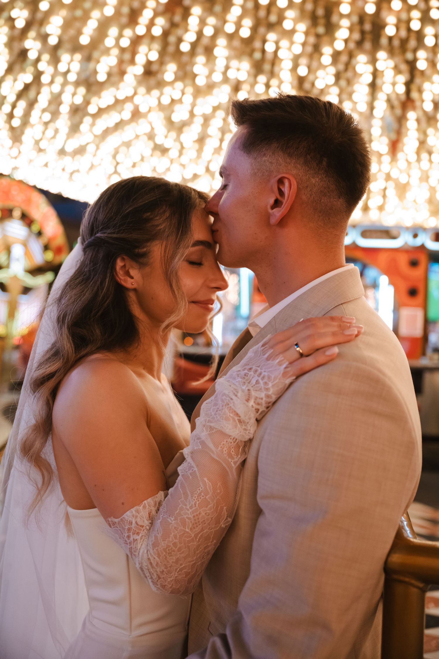 A bride and groom share a romantic moment under warm fairy lights, with the groom kissing her forehead at their wedding reception.