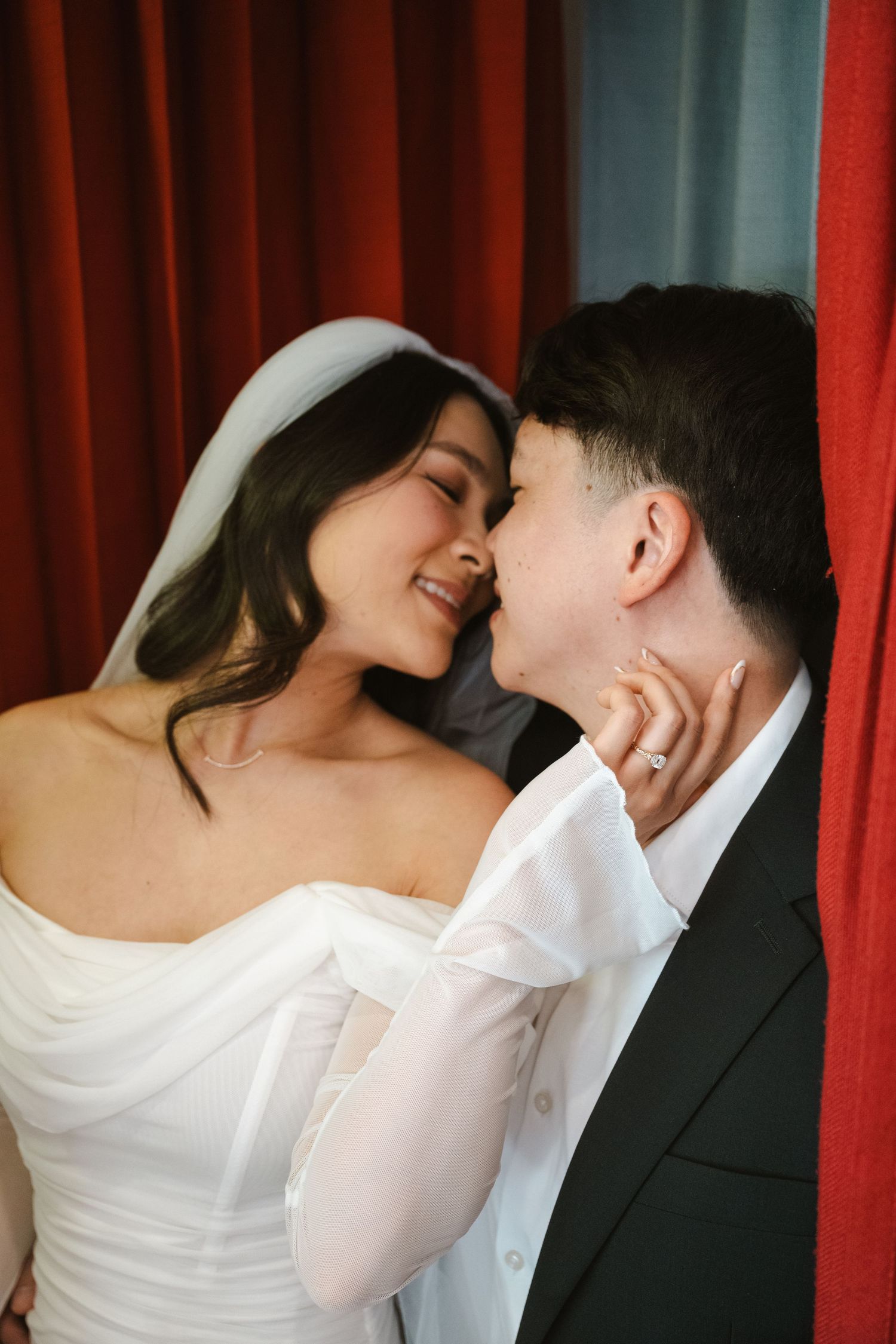 A bride in a white off-shoulder gown and veil shares a romantic moment with her partner in a black suit.