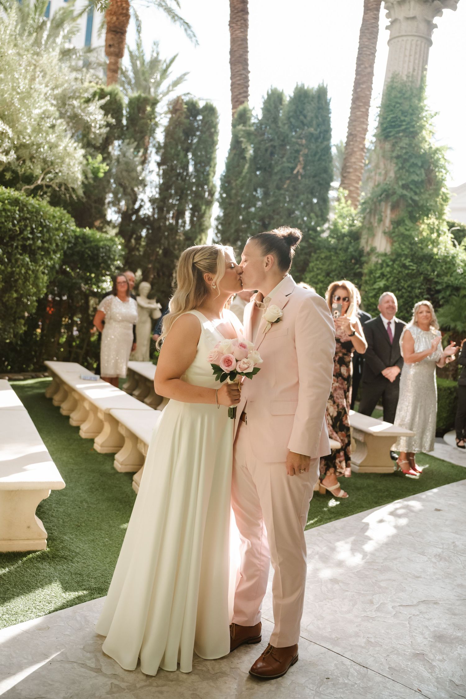 Wedding couple dancing together on a sunny patio surrounded by palm trees and guests at an outdoor reception.