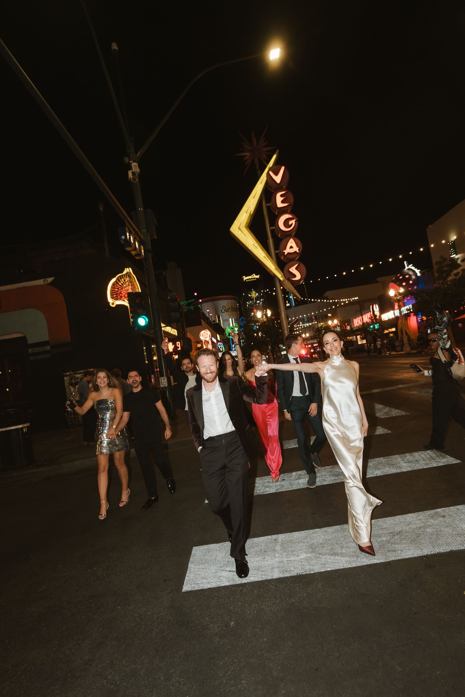 Couple in formal attire crossing Las Vegas street at night with Vegas neon sign glowing above wedding party.