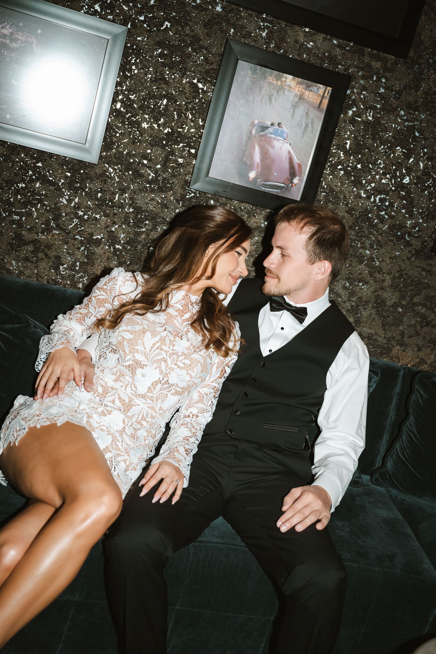 Elegant couple in wedding attire sharing a romantic moment on a velvet sofa in a glamorous venue.
