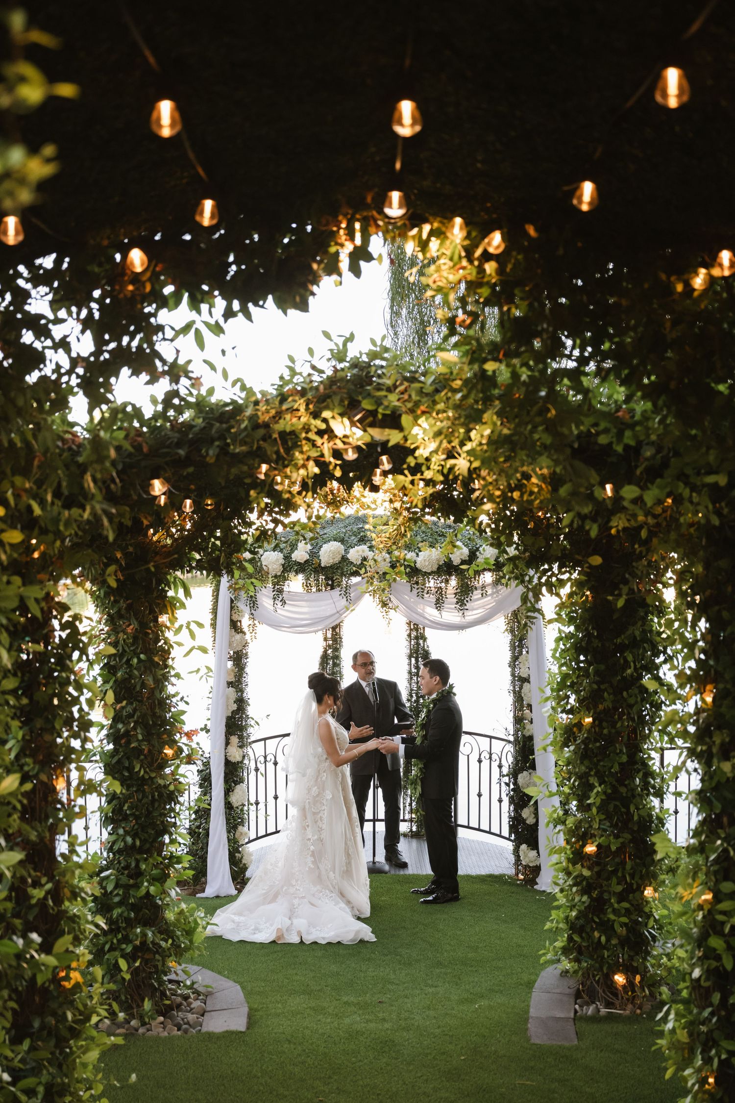 Bride and groom exchange vows under a lush floral arch with string lights at an elegant outdoor wedding ceremony.