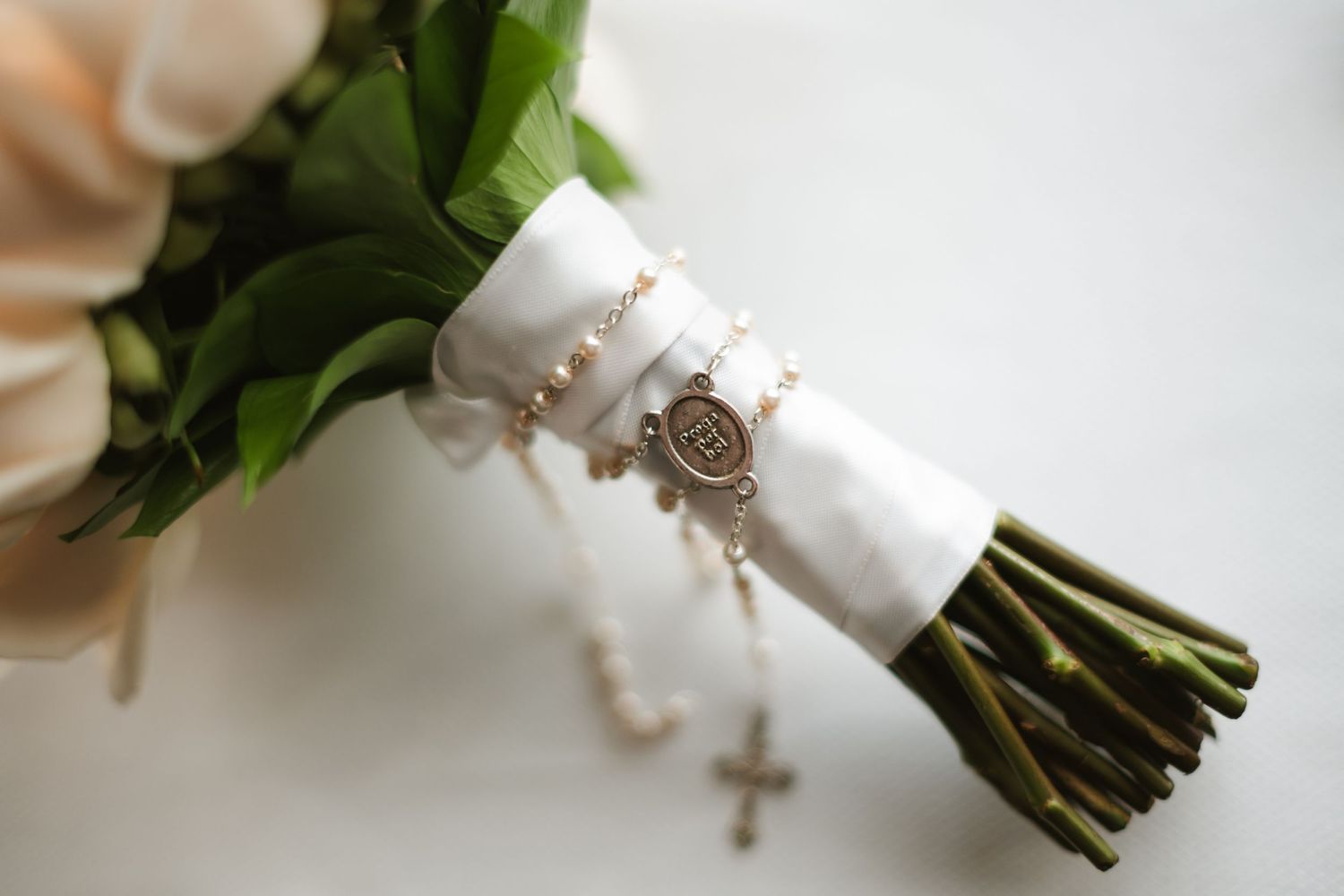 White wedding bouquet with roses and greenery wrapped in satin ribbon and adorned with a rosary.
