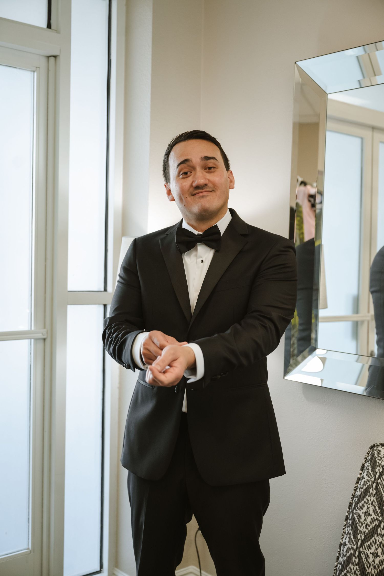 A groom in a black tuxedo with bow tie adjusts his cufflinks while smiling, standing near a window before his wedding.